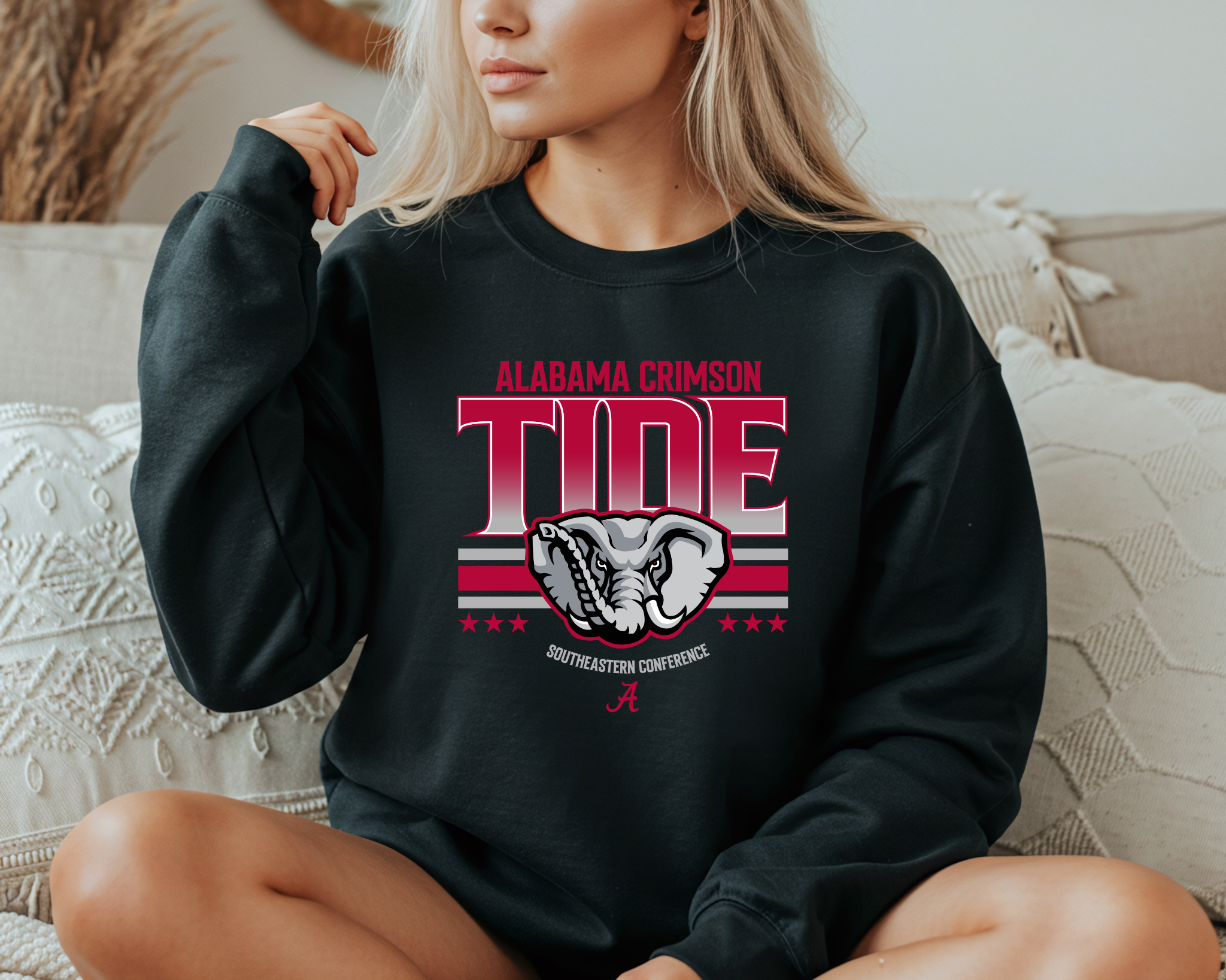 A woman wearing an Alabama Crimson Tide-inspired crewneck sweatshirt in black, featuring bold crimson and gray graphics with an elephant design, sitting comfortably on a couch.