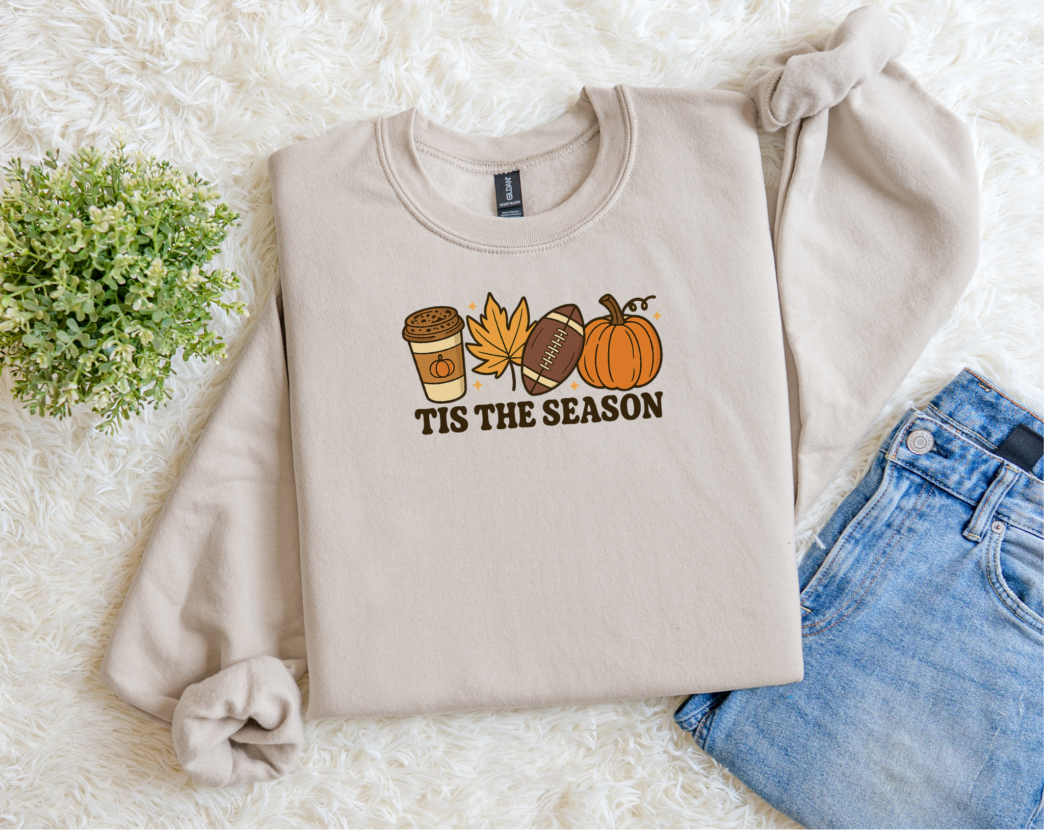 Beige sweatshirt with 'TIS THE SEASON' graphic on a white surface with jeans and flowers.