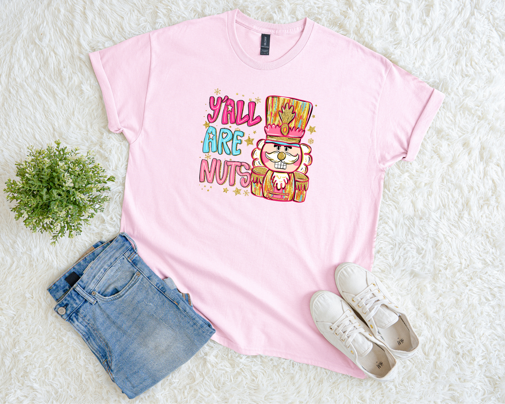 A cream-colored Christmas t-shirt featuring a bright nutcracker design with the phrase “Y’all Are Nuts,” styled flat with jeans and sneakers on a soft white background.