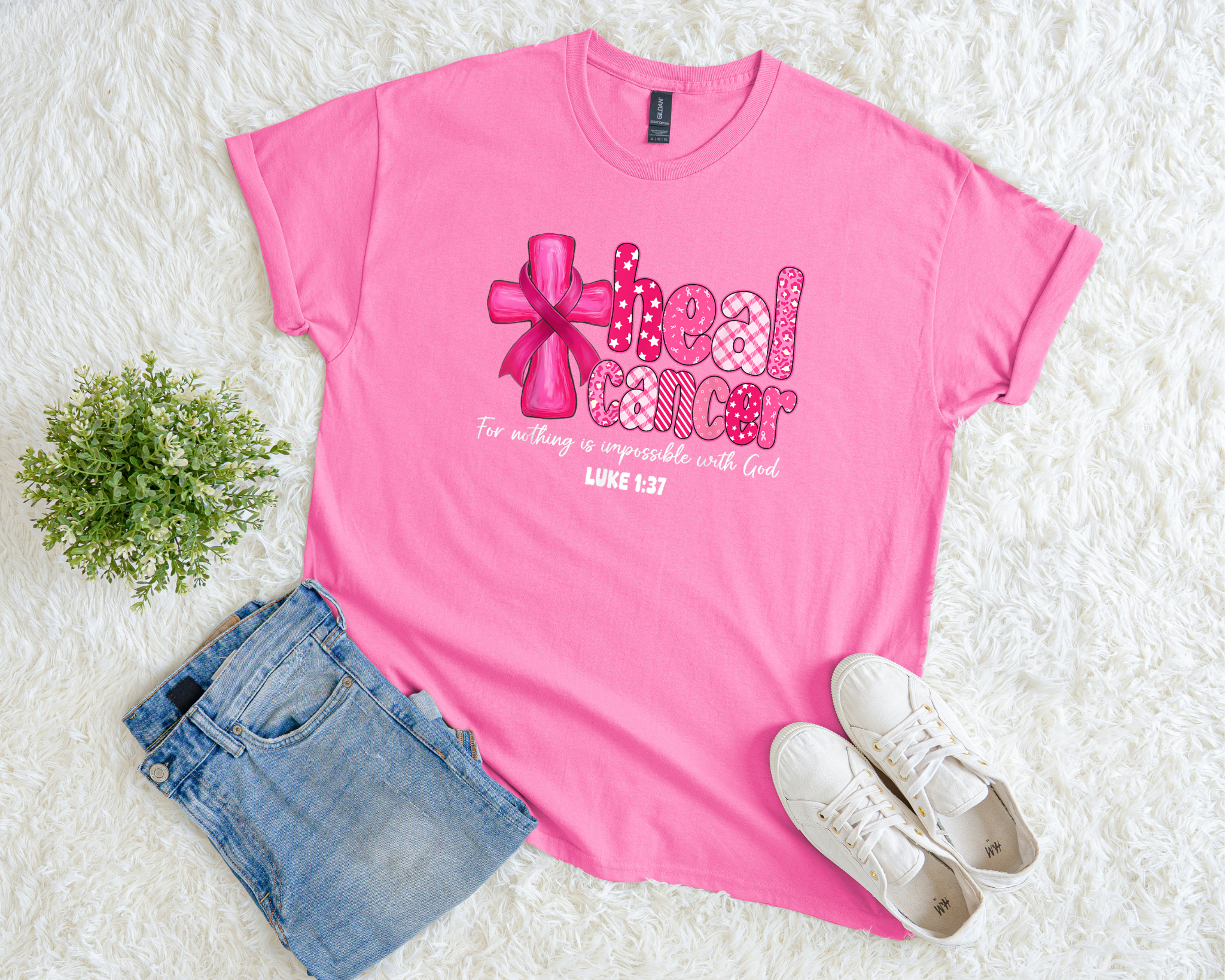 A bright pink t-shirt with a cross and pink ribbon design that reads “Heal Cancer” with Luke 1:37 scripture, styled flat with jeans, white sneakers, and a small green plant on a soft white background.
