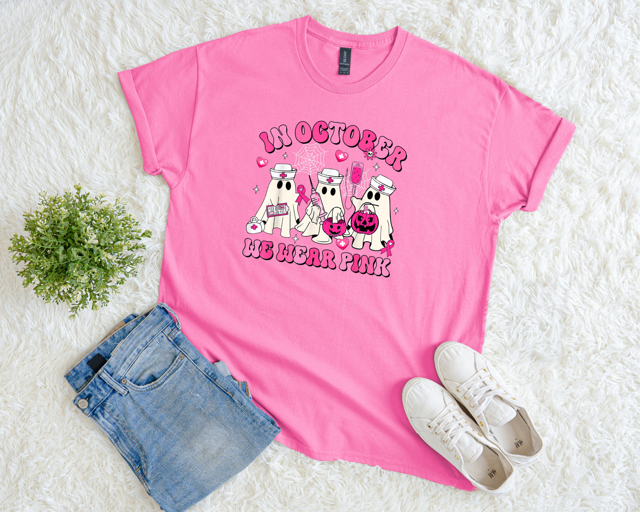 A bright pink t-shirt with cute ghost nurse graphics, pink ribbons, and Halloween accents that read “In October We Wear Pink,” styled flat with jeans, white sneakers, and a small green plant on a fluffy white background.
