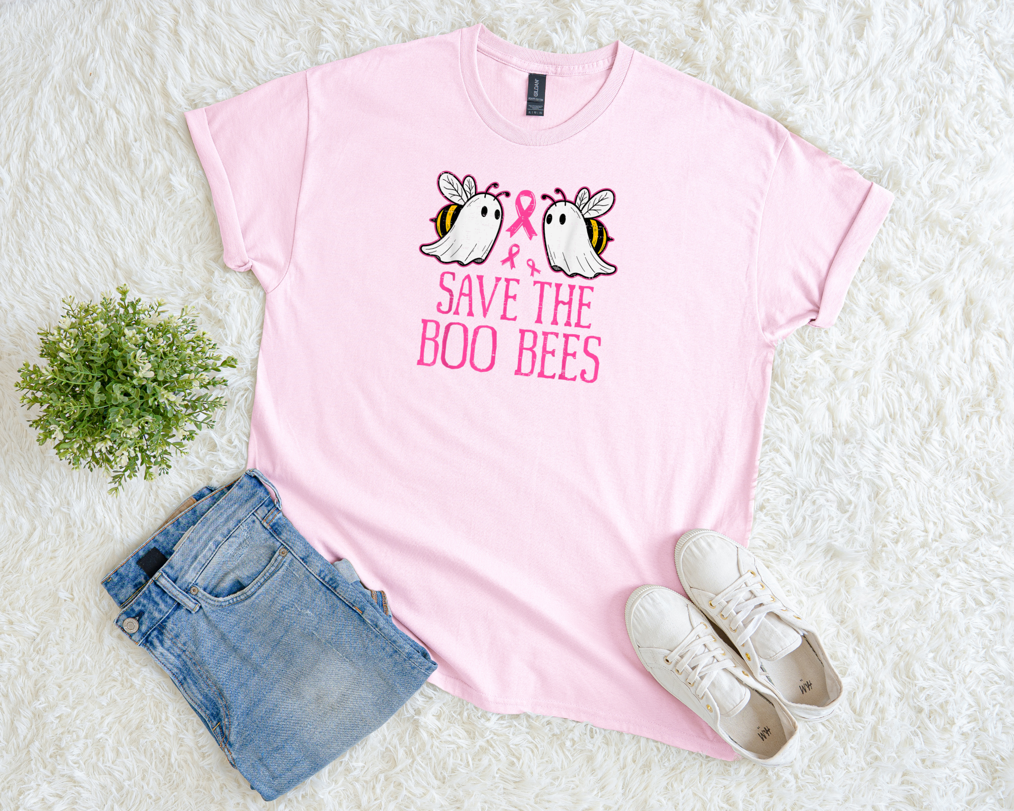 A light pink t-shirt with playful ghost-bee graphics and bold pink text reading “Save the Boo Bees” with a pink ribbon, styled flat with jeans, white sneakers, and a small green plant on a soft white background.