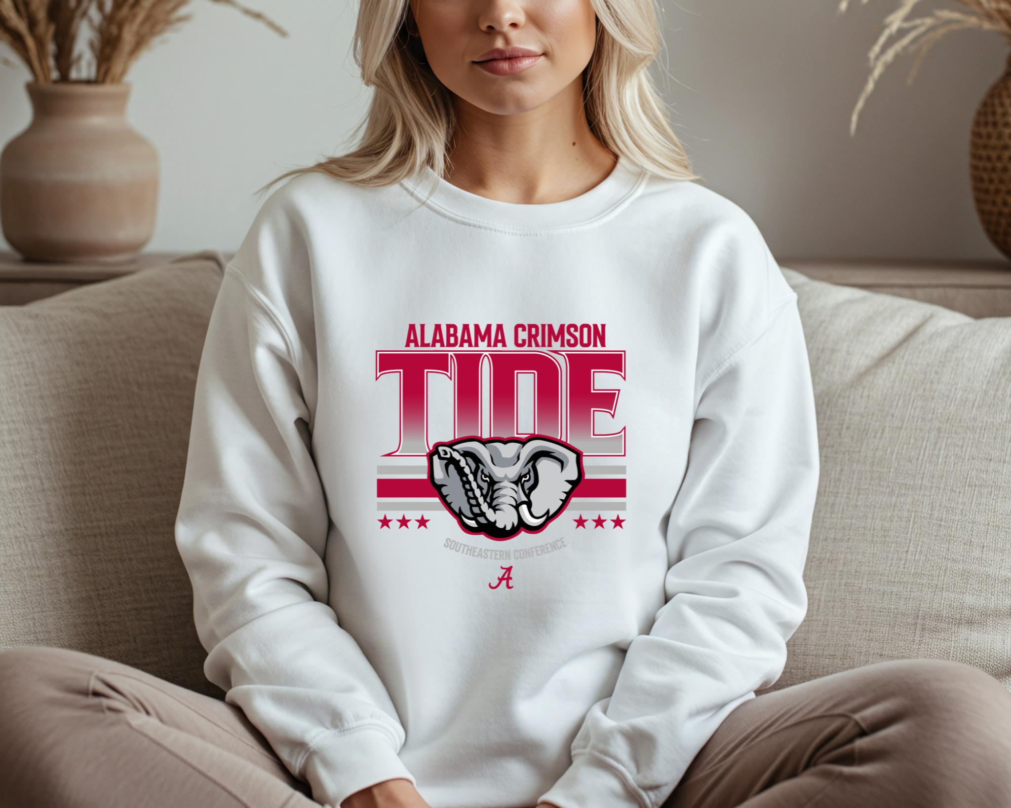A woman wearing an Alabama Crimson Tide-inspired crewneck sweatshirt in black, featuring bold crimson and gray graphics with an elephant design, sitting comfortably on a couch.