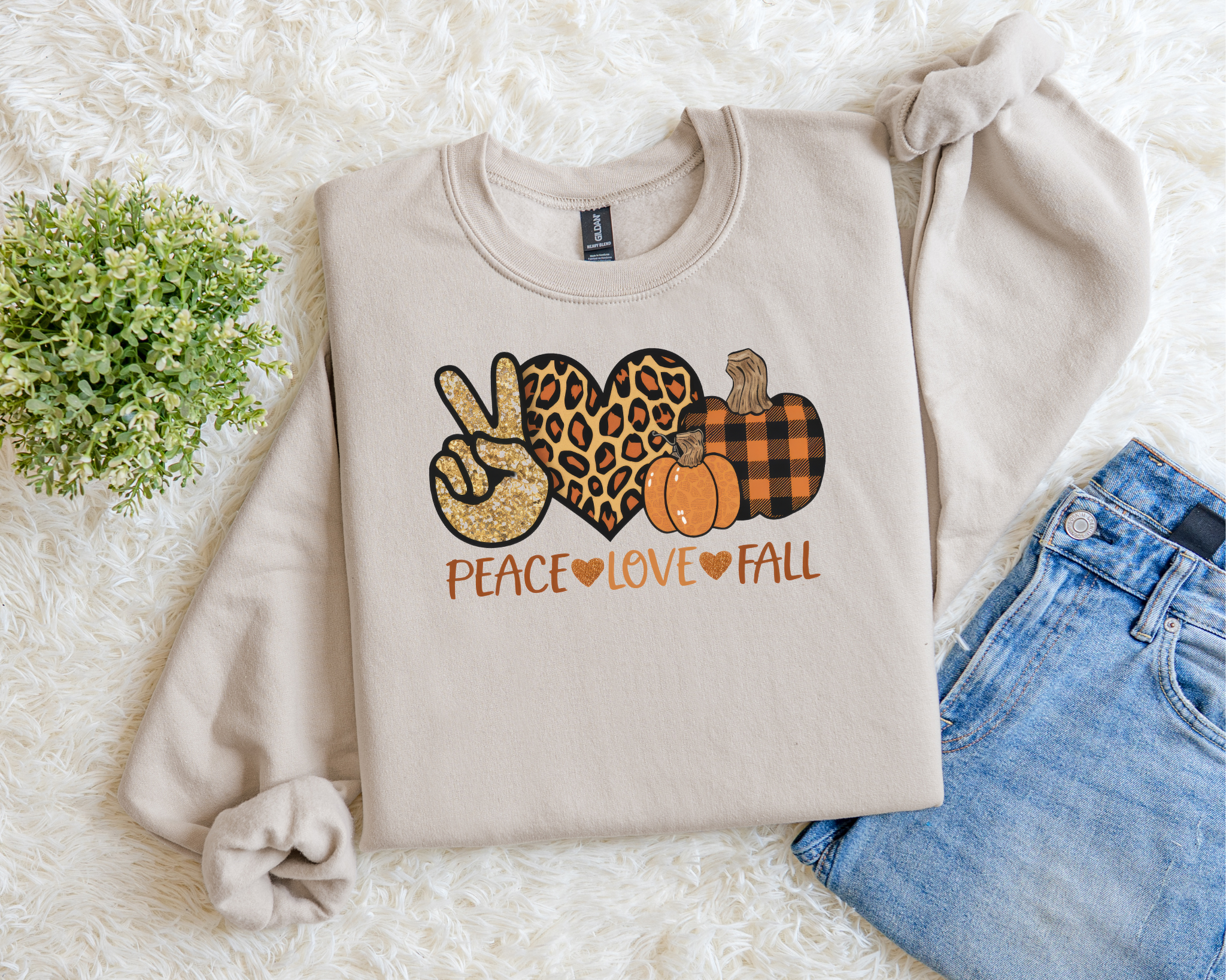 Beige sweatshirt with 'Peace Love Fall' design on a white surface with jeans and greenery.