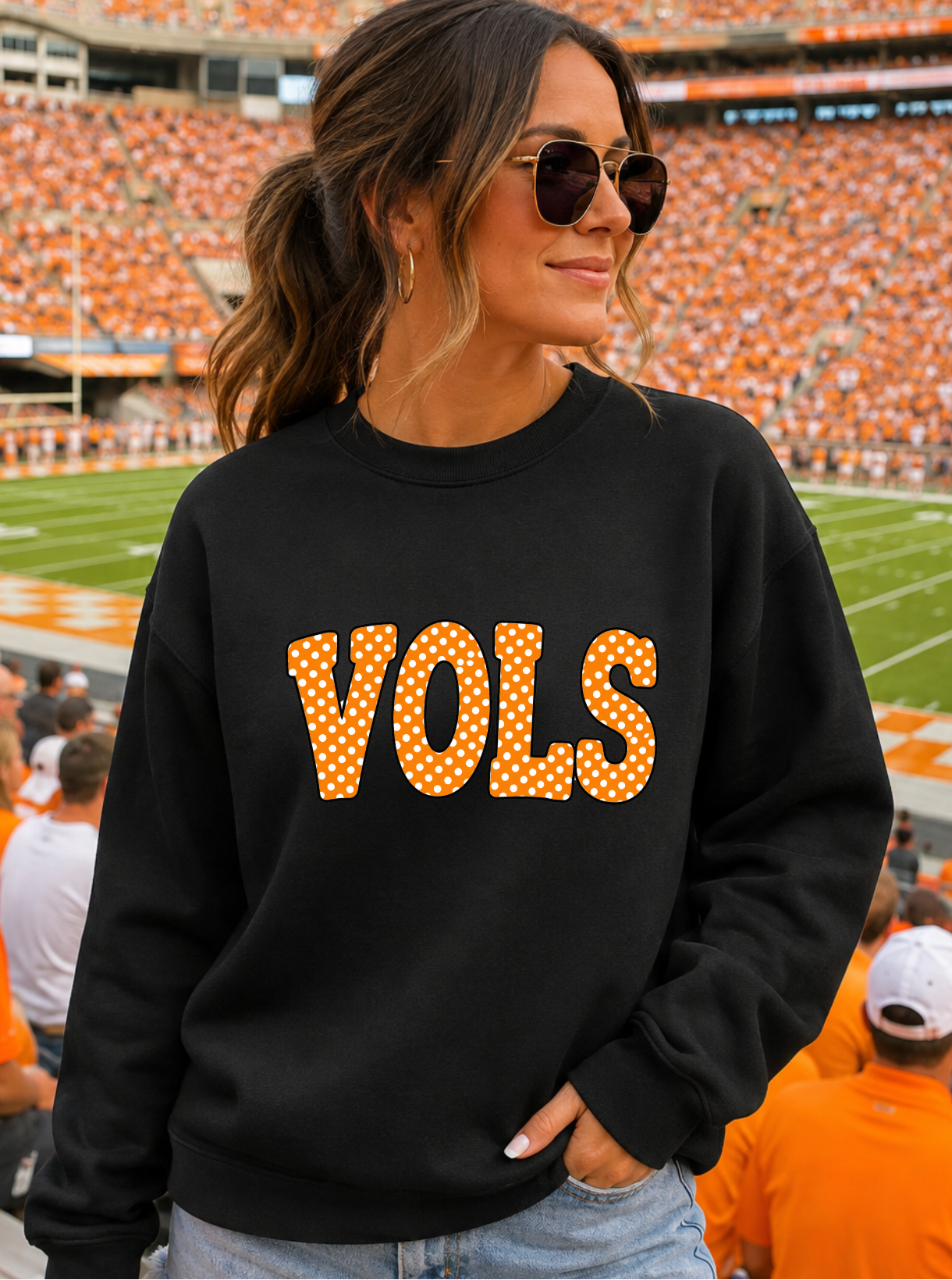 A woman wearing a black crewneck sweatshirt with 'VOLS' in orange letters outlined with white dots, standing in a stadium with orange seats in the background.