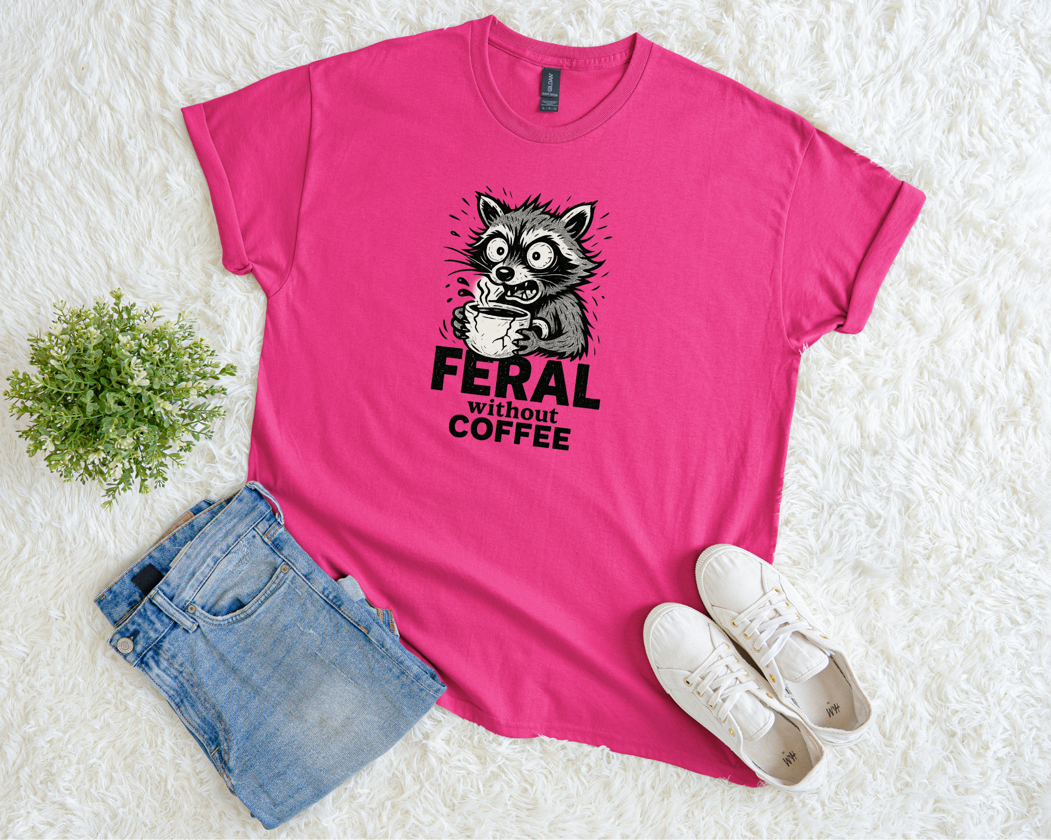 Pink t-shirt with a graphic of a raccoon holding coffee and text 'Feral without coffee' on a white surface with jeans and shoes.