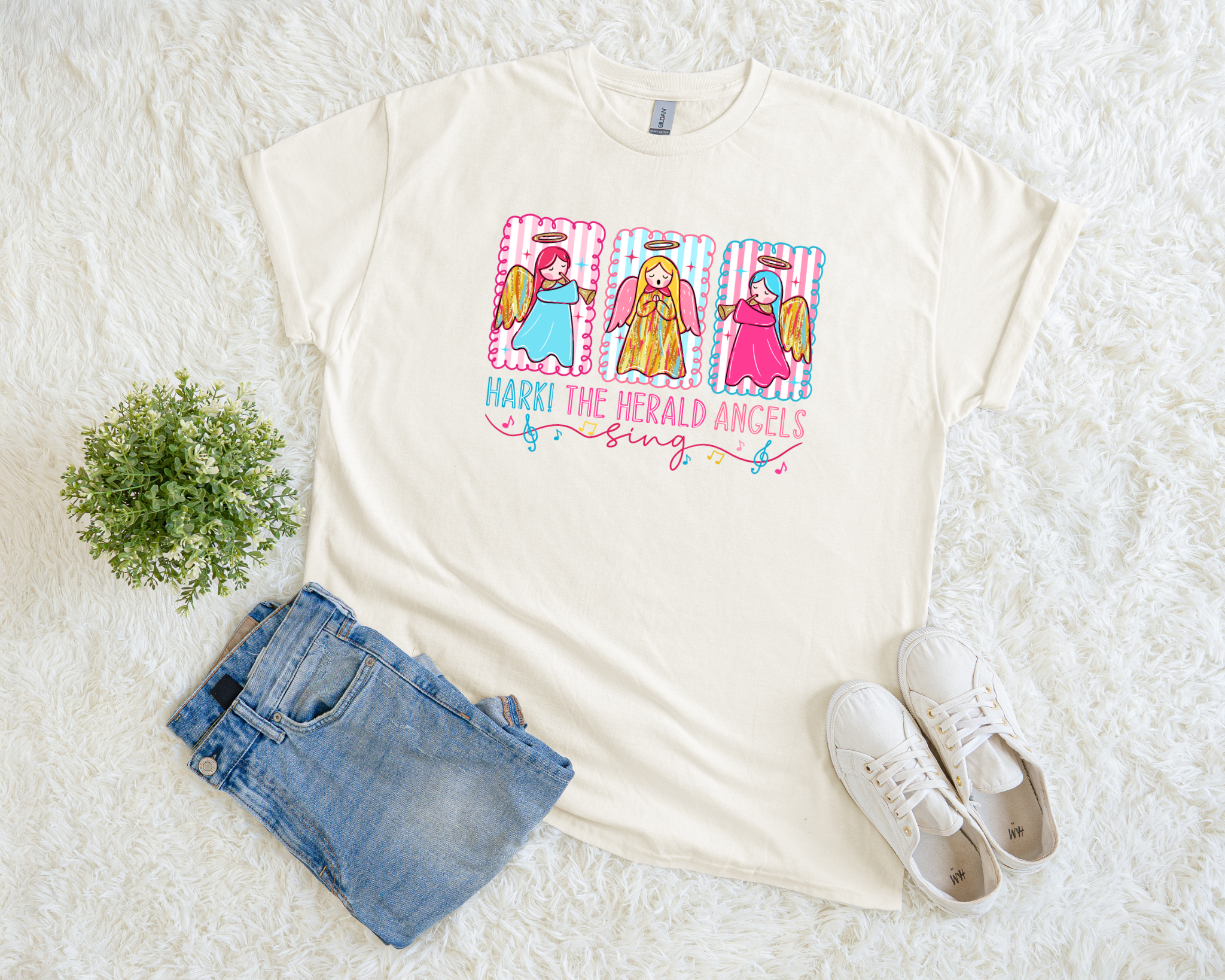 A cream-colored Christmas t-shirt featuring three colorful angels with gold wings and the phrase “Hark! The Herald Angels Sing,” styled flat with jeans and sneakers on a soft white background.