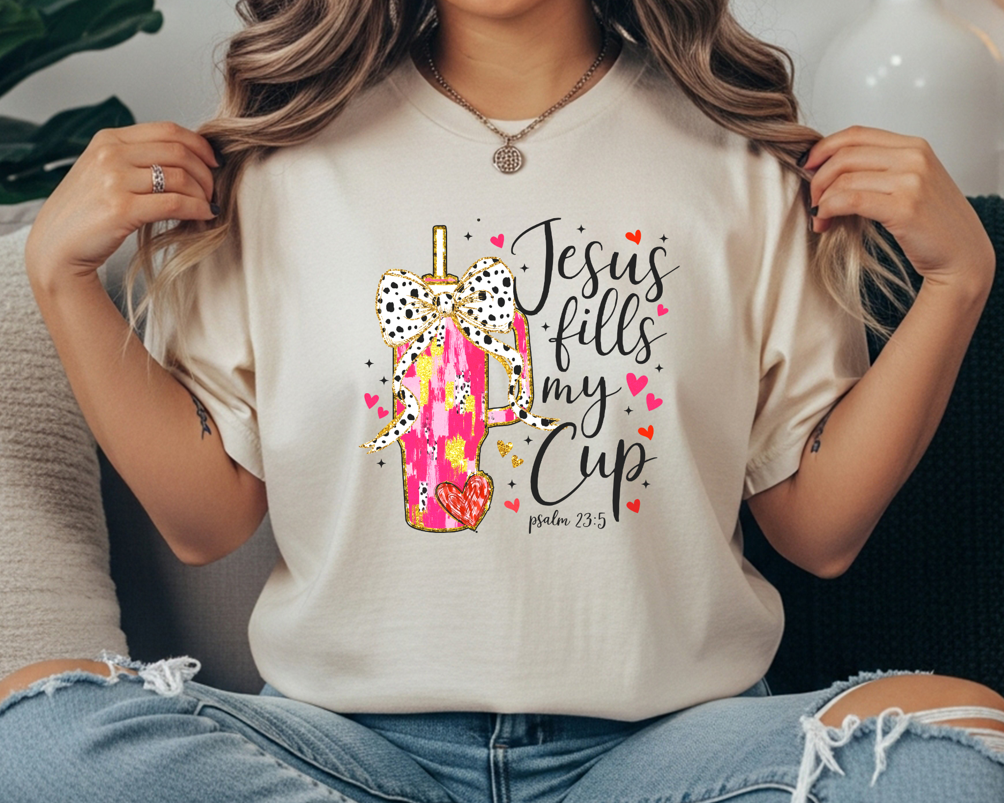 Comfort Colors cream t-shirt featuring a pink tumbler with a bow graphic and the phrase “Jesus fills my cup” from Psalm 23:5, styled with ripped jeans and neutral accessories.