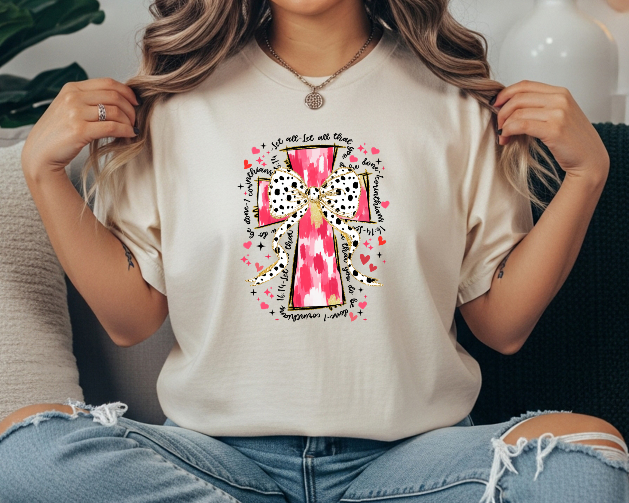 Comfort Colors ivory t-shirt featuring a pink watercolor cross with a black and white polka dot bow, surrounded by hand-lettered faith scripture text and pink heart accents, styled with ripped jeans in a cozy lifestyle flat lay