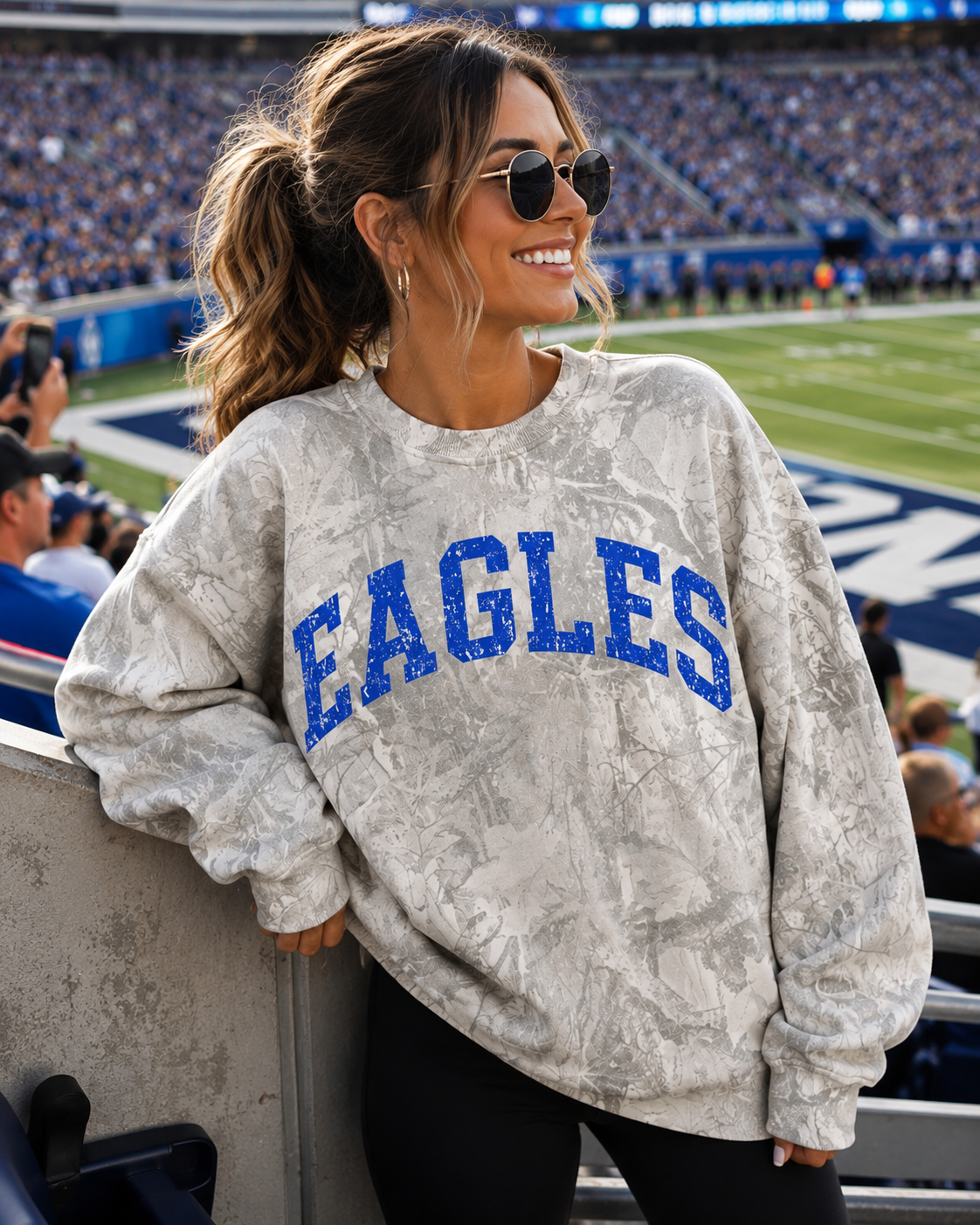 A woman in a stadium wearing a light gray marbled sweatshirt with 'EAGLES' printed in blue across the chest, paired with black pants and sunglasses.