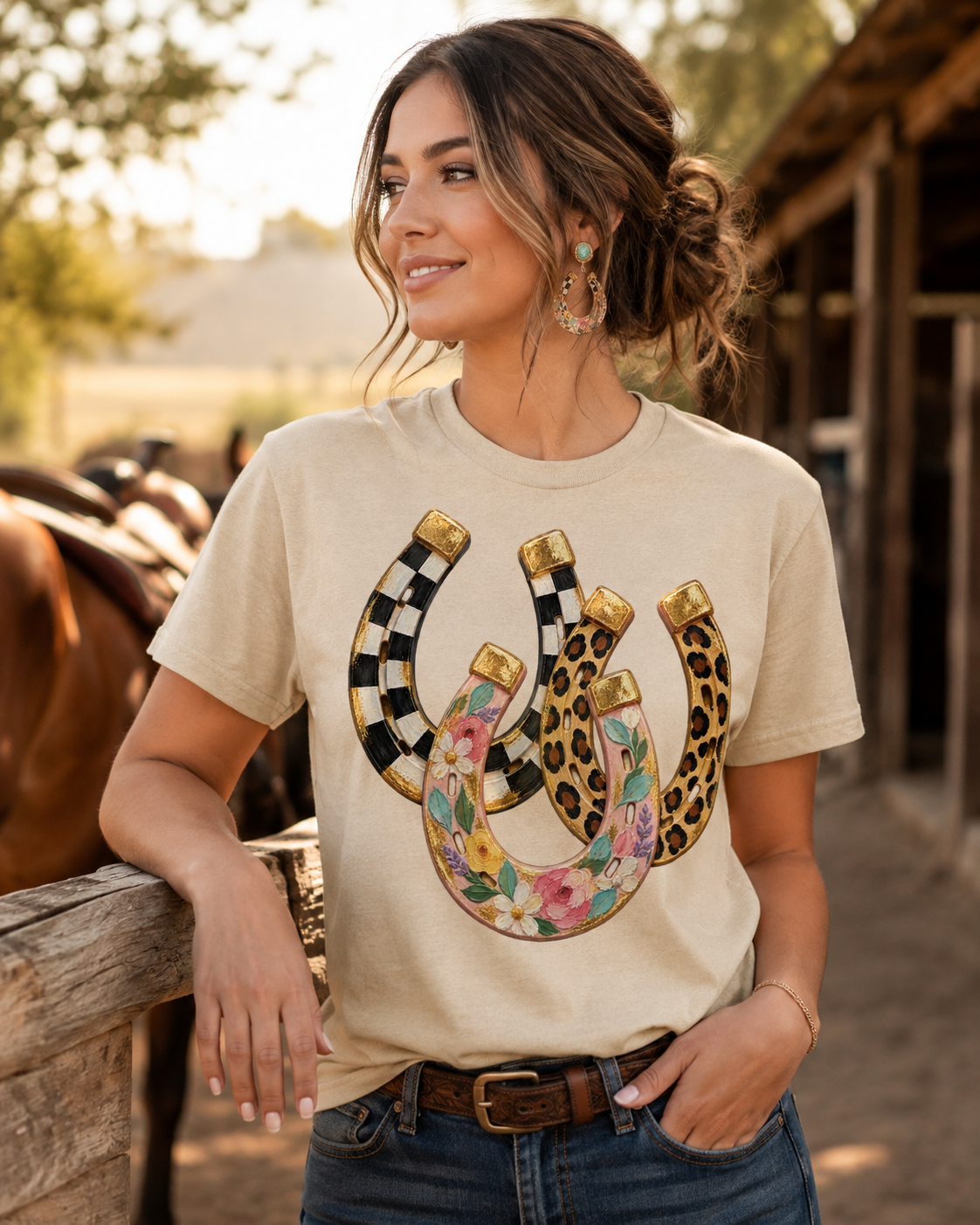 A woman wearing a beige graphic t-shirt with three stylized horseshoes (black-and-white checkered, leopard print, and floral) paired with blue jeans in an outdoor rustic barn setting.