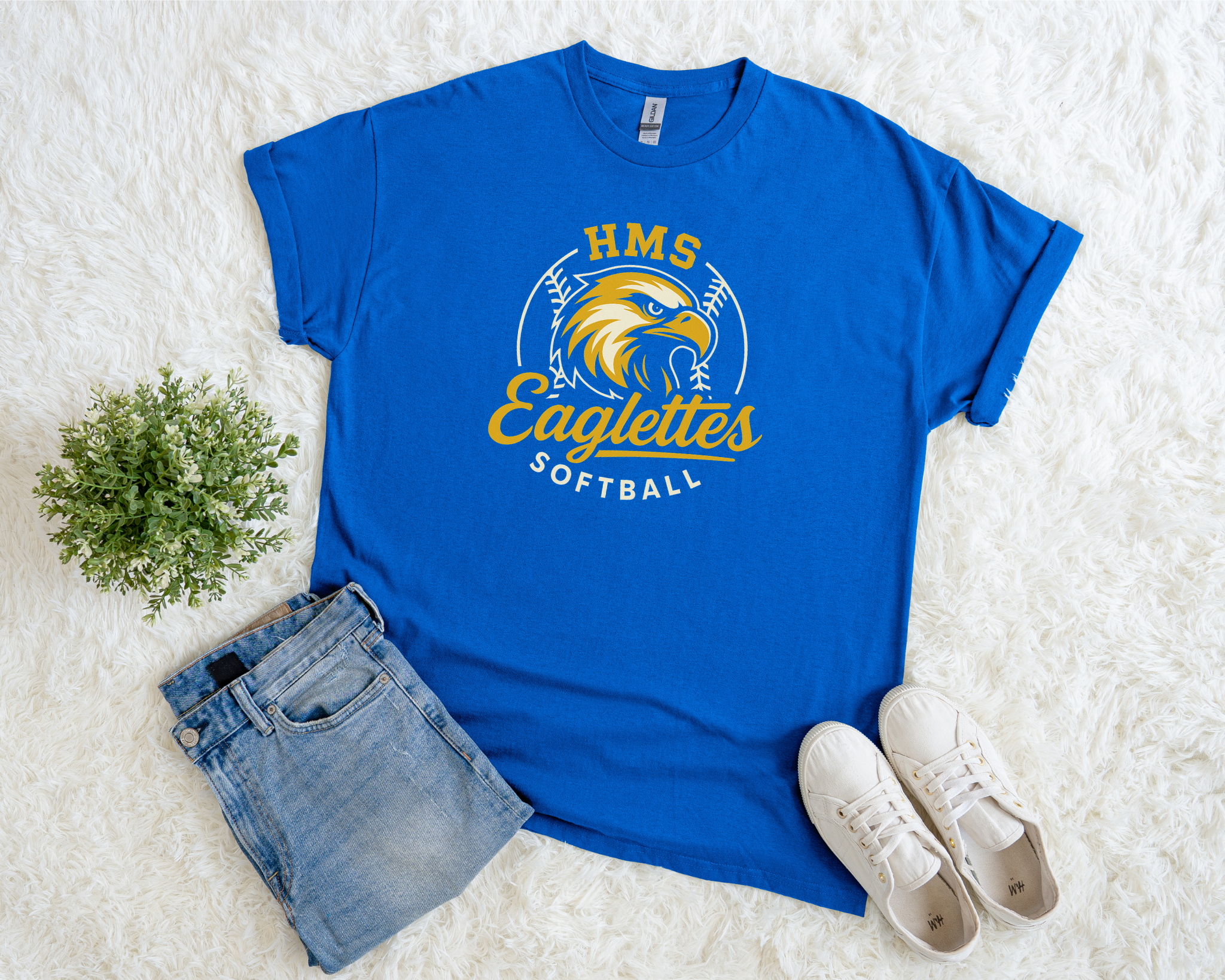 A royal blue HMS Eaglettes Softball t-shirt with a yellow eagle graphic and team name, styled flat with jeans, white sneakers, and a green plant on a soft white background.