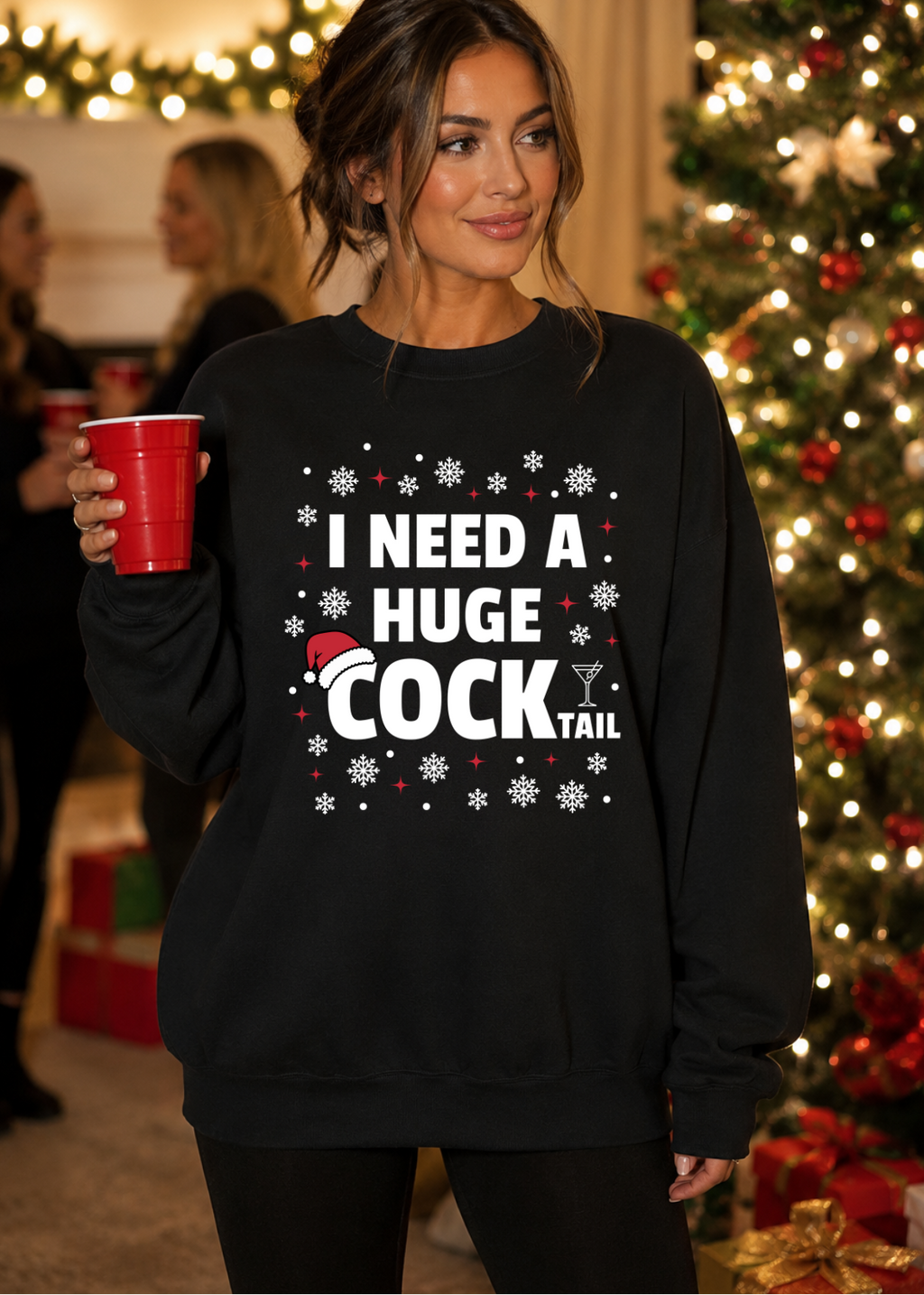 A woman wearing a black crewneck sweatshirt with a white graphic that reads 'I NEED A HUGE COCKTAIL' surrounded by snowflakes and a Santa hat, holding a red cup in front of a decorated Christmas tree.
