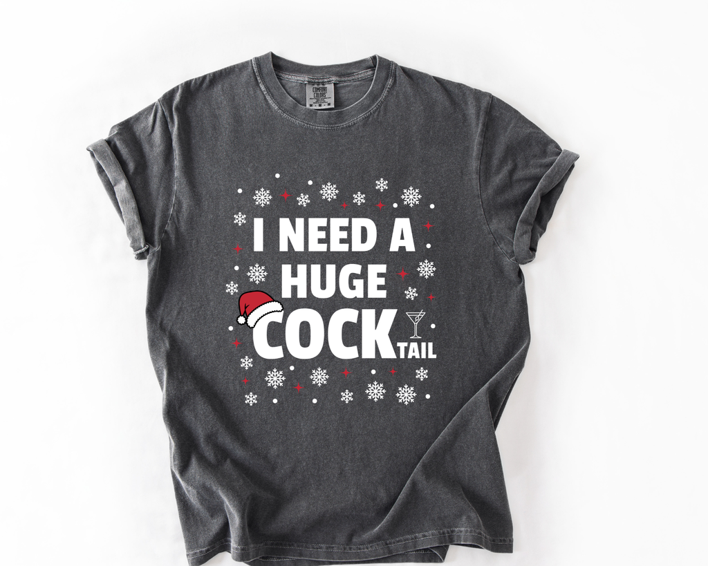 A dark gray t-shirt with a humorous graphic that reads 'I NEED A HUGE COCK TAIL' in white text, decorated with white snowflakes and a small red and white Santa hat, laid flat on a white background.