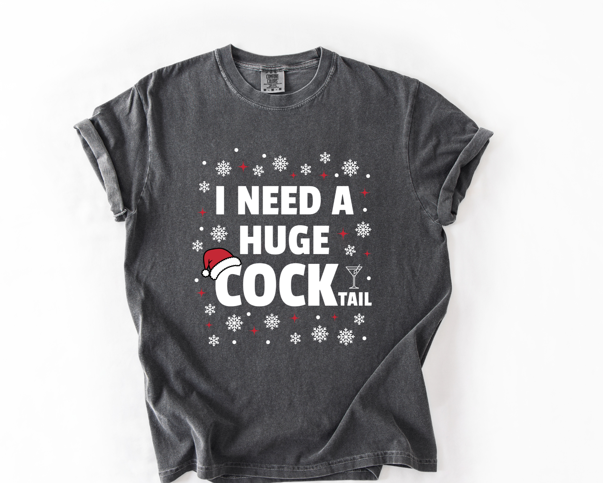 A dark gray t-shirt with a humorous graphic that reads 'I NEED A HUGE COCK TAIL' in white text, decorated with white snowflakes and a small red and white Santa hat, laid flat on a white background.