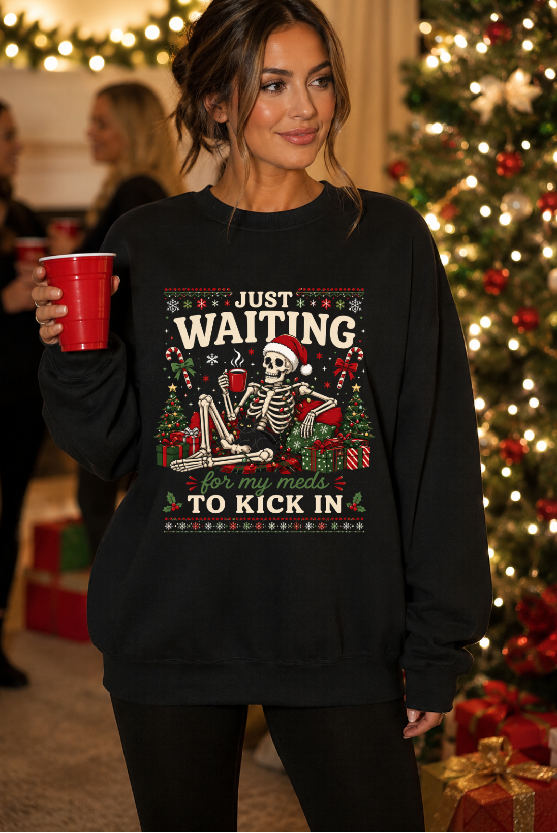 A woman wearing a black crewneck sweatshirt with a festive Christmas graphic that reads 'JUST WAITING for my man to KICK IN'. She is holding a red cup in a decorated holiday home with a Christmas tree in the background.