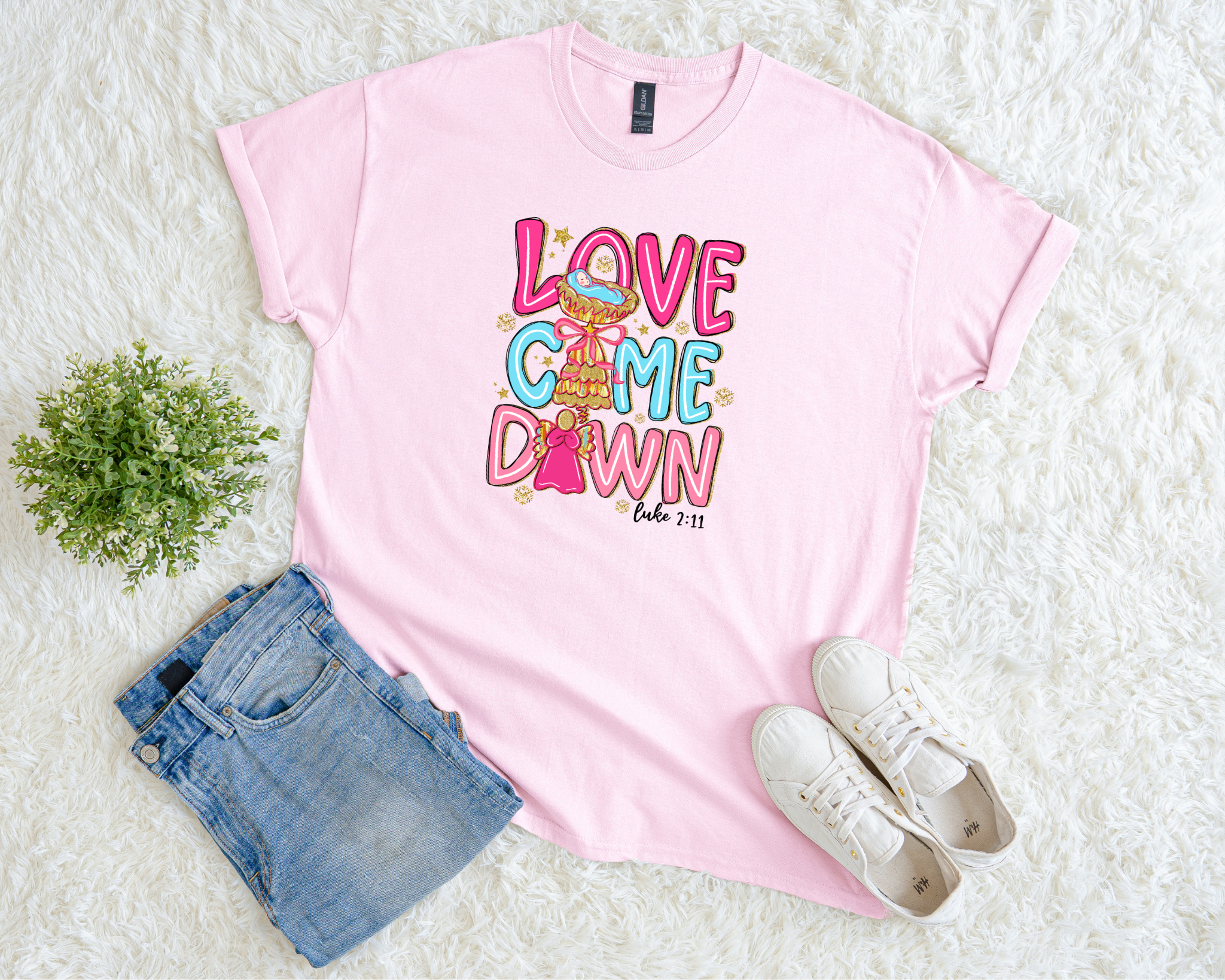 Light pink t-shirt with colorful “Love Came Down” text and Luke 2:11 Bible verse, styled flat with jeans, sneakers, and a plant on a soft white background.
