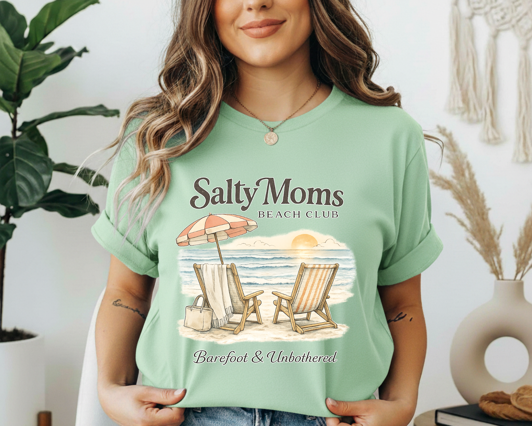 Woman wearing a green t-shirt with 'Salty Moms Beach Club' design, standing indoors.