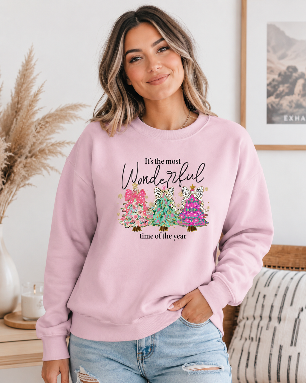 A woman wearing a light pink crewneck sweatshirt with a graphic print that reads 'It's the most Wonderful time of the year' above three colorful Christmas trees. She is posing in a cozy, brightly lit room with pampas grass and a striped cushion.
