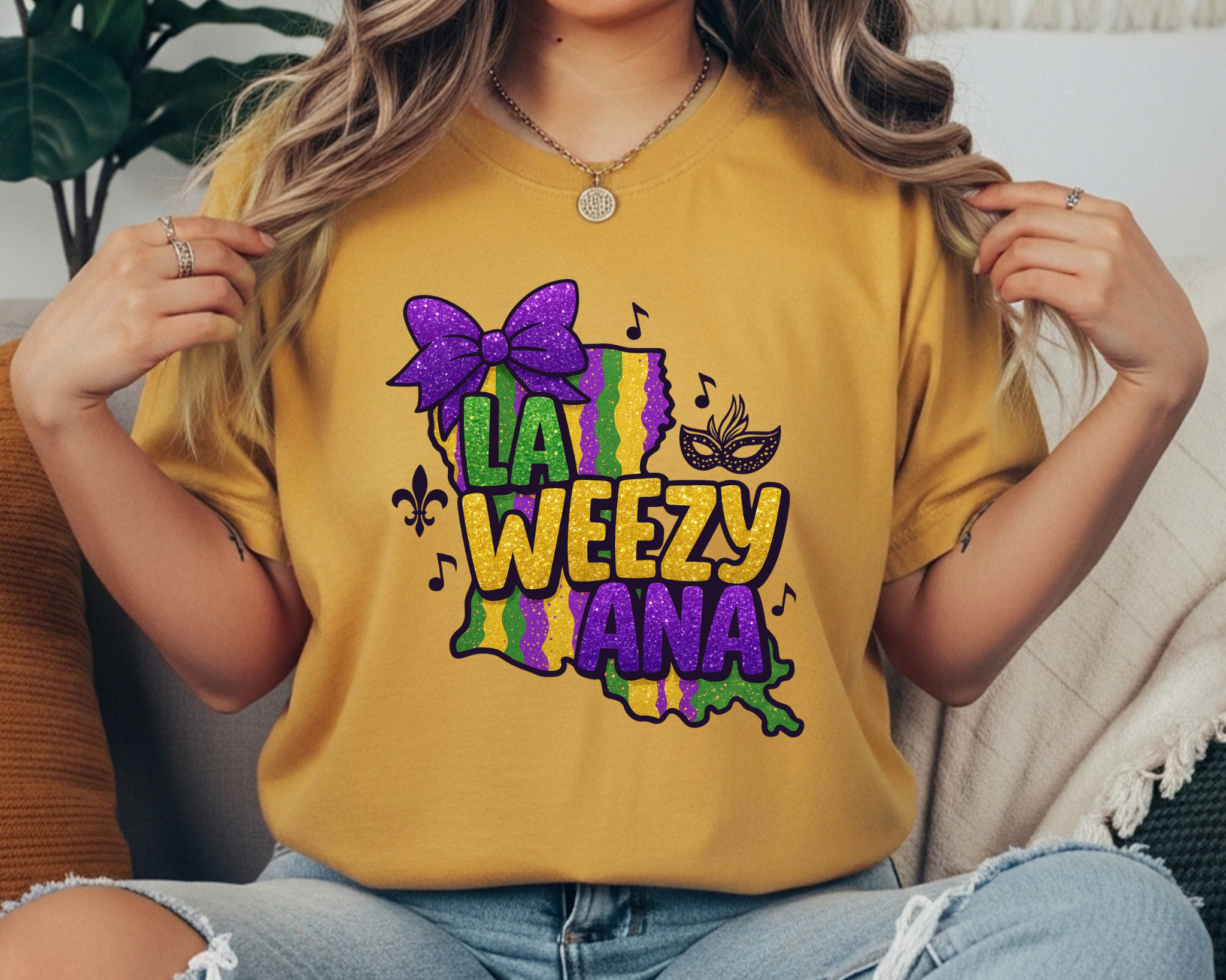La Weezy Ana Mardi Gras t-shirt with a faux glitter-style Louisiana state graphic, purple bow, mask, music notes, and fleur-de-lis in purple, green, and gold