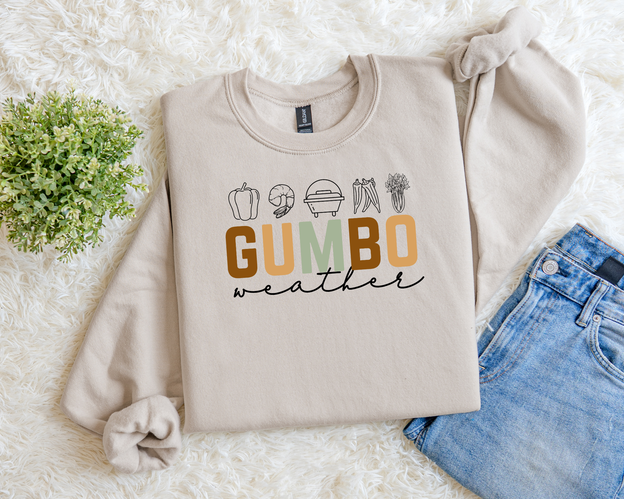 Beige sweatshirt with 'GUMBO weather' text on a white surface with flowers and jeans.