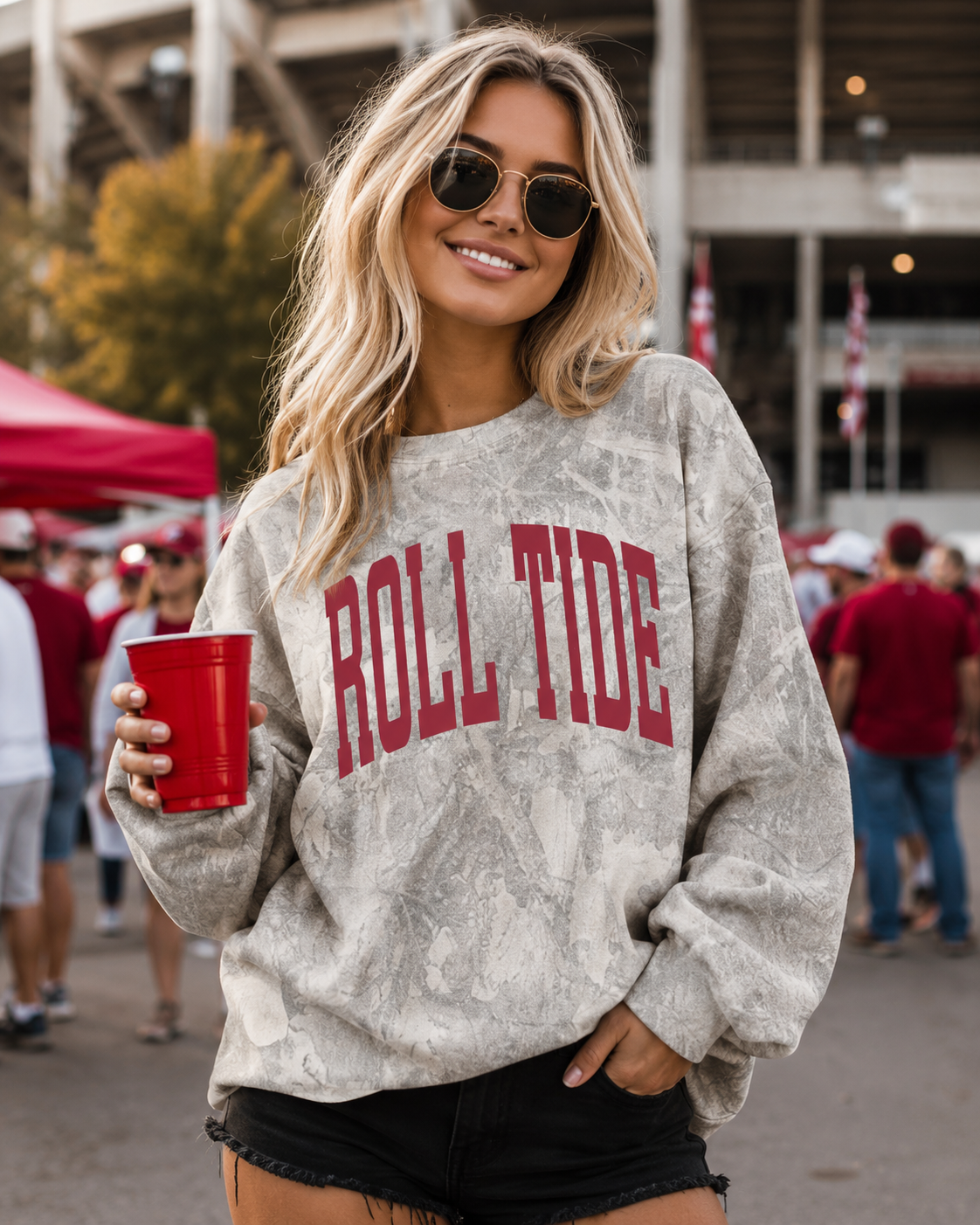 A woman wearing a light gray tie-dye sweatshirt with 'ROLL TIDE' printed in red across the chest, holding a red cup at what appears to be a stadium or event.