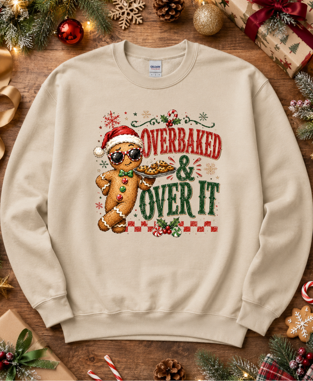 A beige crewneck sweatshirt with a festive Christmas graphic featuring a gingerbread man wearing sunglasses and a Santa hat, with the text 'OVERBAKED & OVER IT' in red and green. The sweatshirt is displayed in a flat-lay arrangement on a rustic wooden surface surrounded by holiday decorations like pine branches, ornaments, and wrapped gifts.