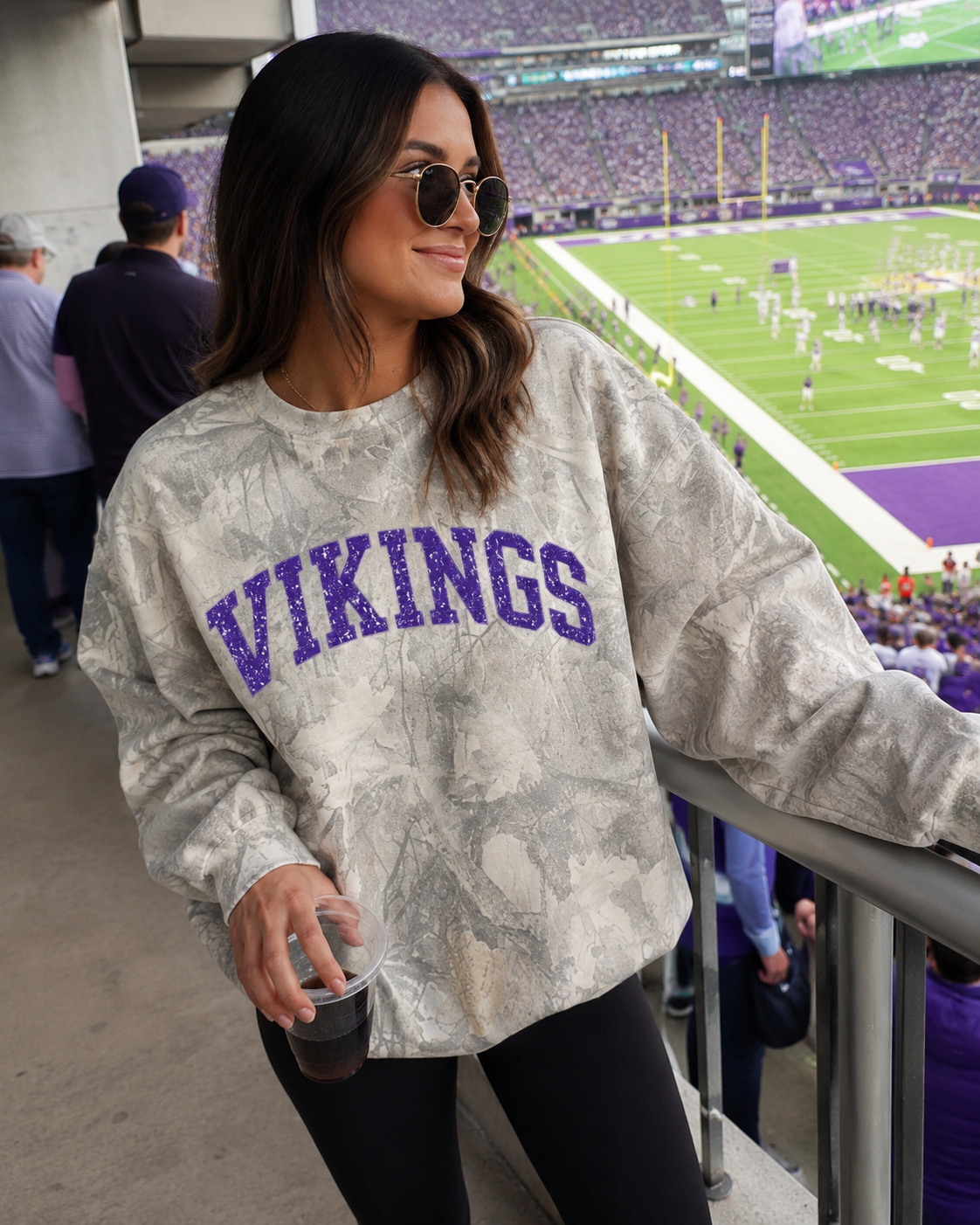 A woman in a stadium stands wearing a gray and white tie-dye sweatshirt with 'VIKINGS' in purple across the chest, paired with black pants. The background shows a football field and crowd.