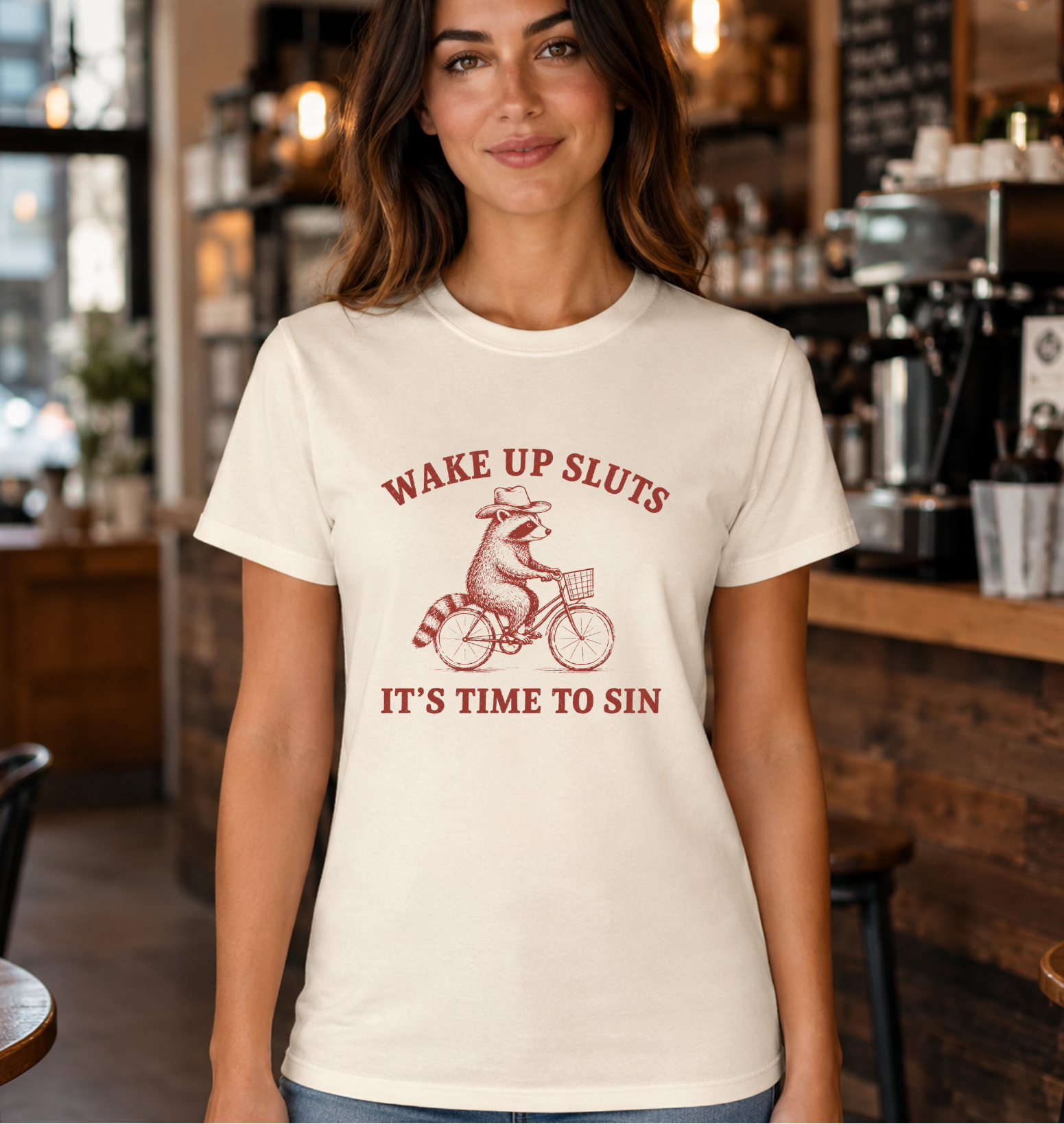 A woman wearing a cream-colored t-shirt with a red graphic of a raccoon on a bicycle and the text 'WAKE UP SLUTS IT'S TIME TO SIN', standing in a coffee shop.