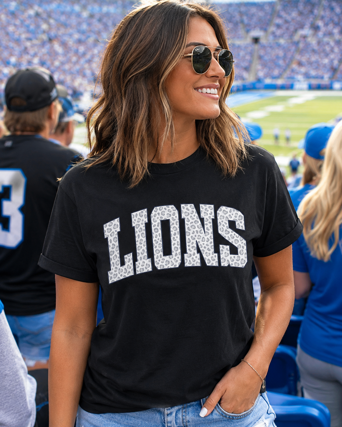 A woman at a stadium wearing a black crewneck t-shirt with 'LIONS' printed across the front in a textured white pattern, paired with light blue jeans and sunglasses.