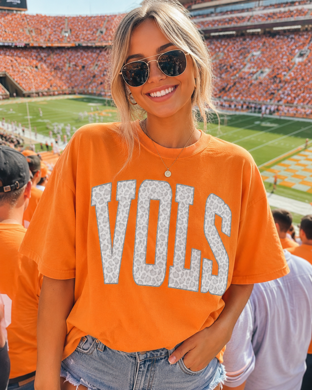 A woman wearing an orange short-sleeve t-shirt with 'VOLS' printed in large white letters, standing in a crowded football stadium.