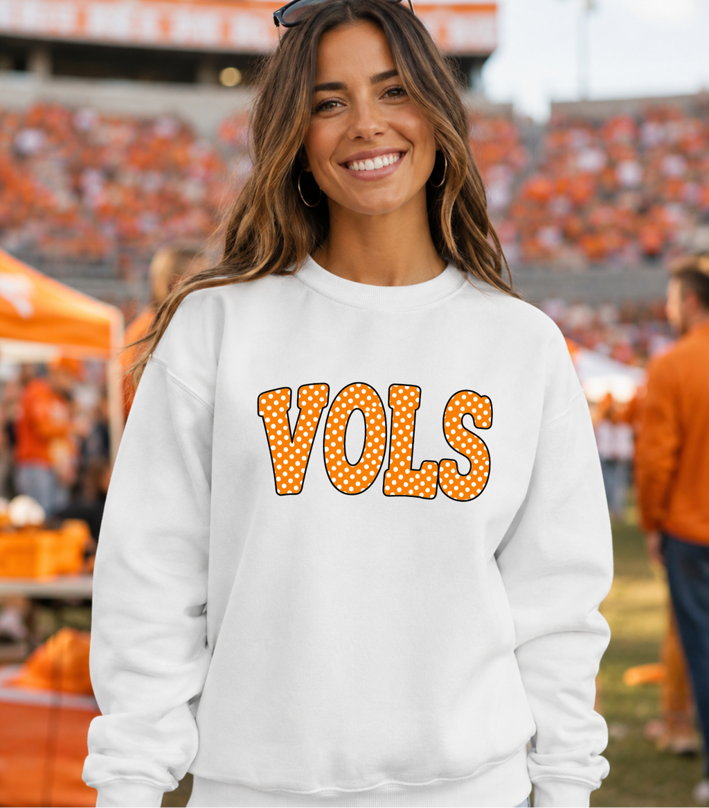 A smiling woman wearing a white crewneck sweatshirt with 'VOLS' printed across the chest in orange letters filled with small white dots and outlined in black. The background shows a blurred outdoor stadium scene with orange seating and people.