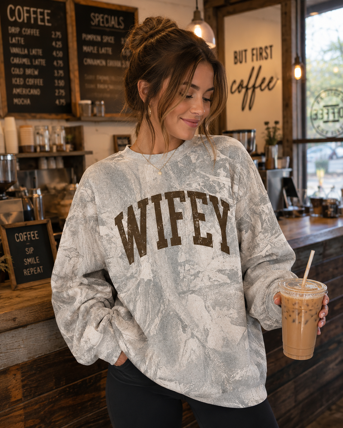 A woman in a coffee shop wearing a light gray and white marbled sweatshirt with 'WIFEY' printed in brown across the chest, holding an iced coffee.