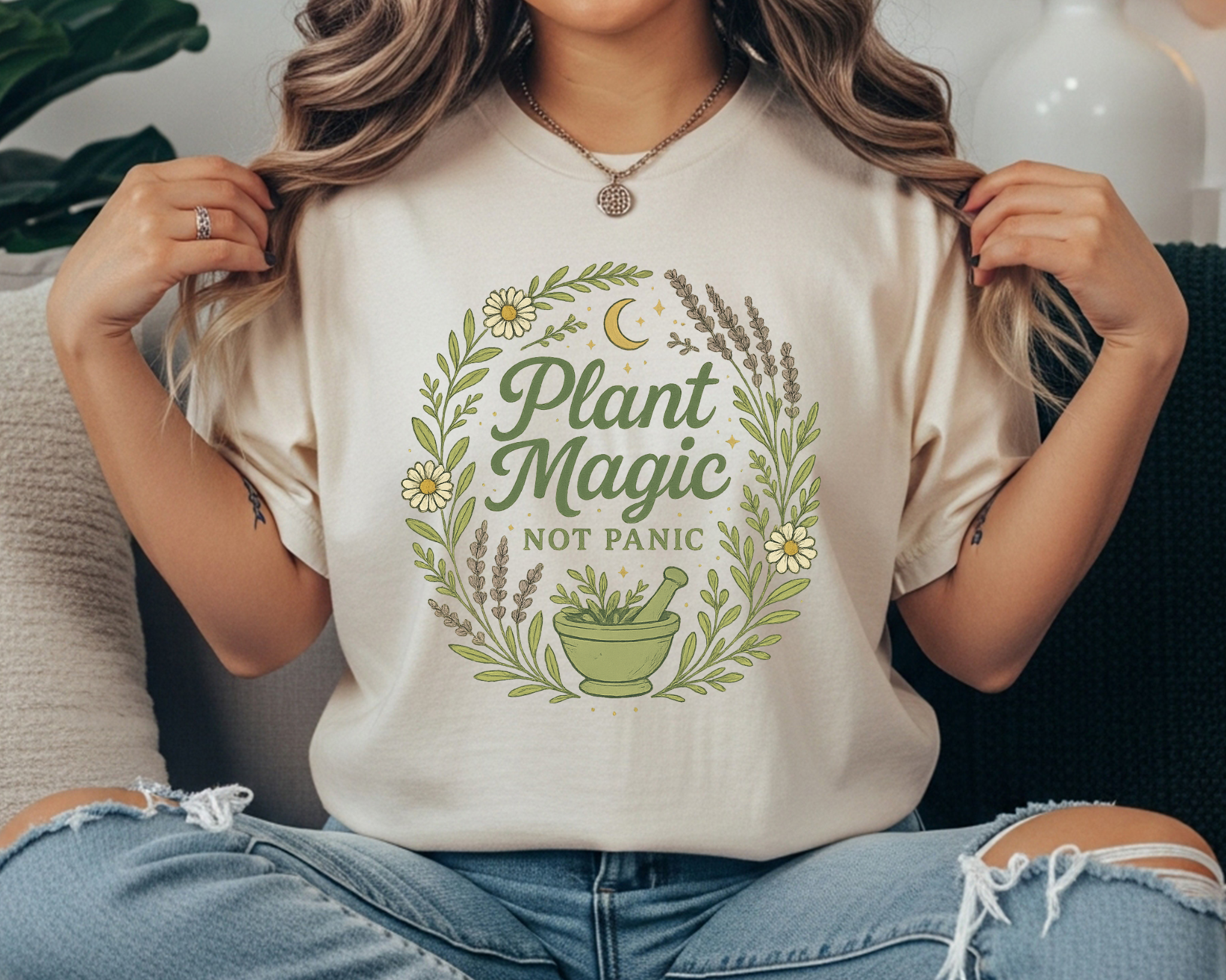 Woman wearing a neutral Comfort Colors t-shirt with a circular wreath of herbs and flowers, a green mortar and pestle, crescent moon, and the text “Plant Magic Not Panic.”