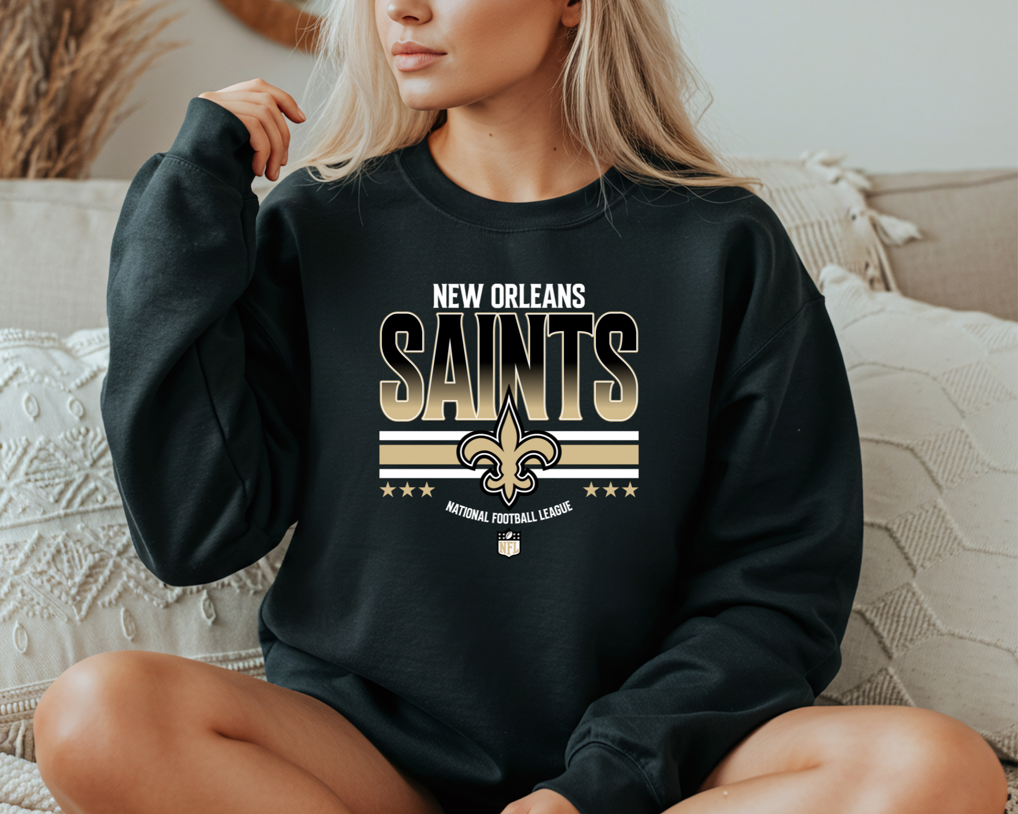A woman wearing a black New Orleans Saints-inspired crewneck sweatshirt featuring bold gold lettering and a fleur-de-lis design, sitting comfortably on a couch.