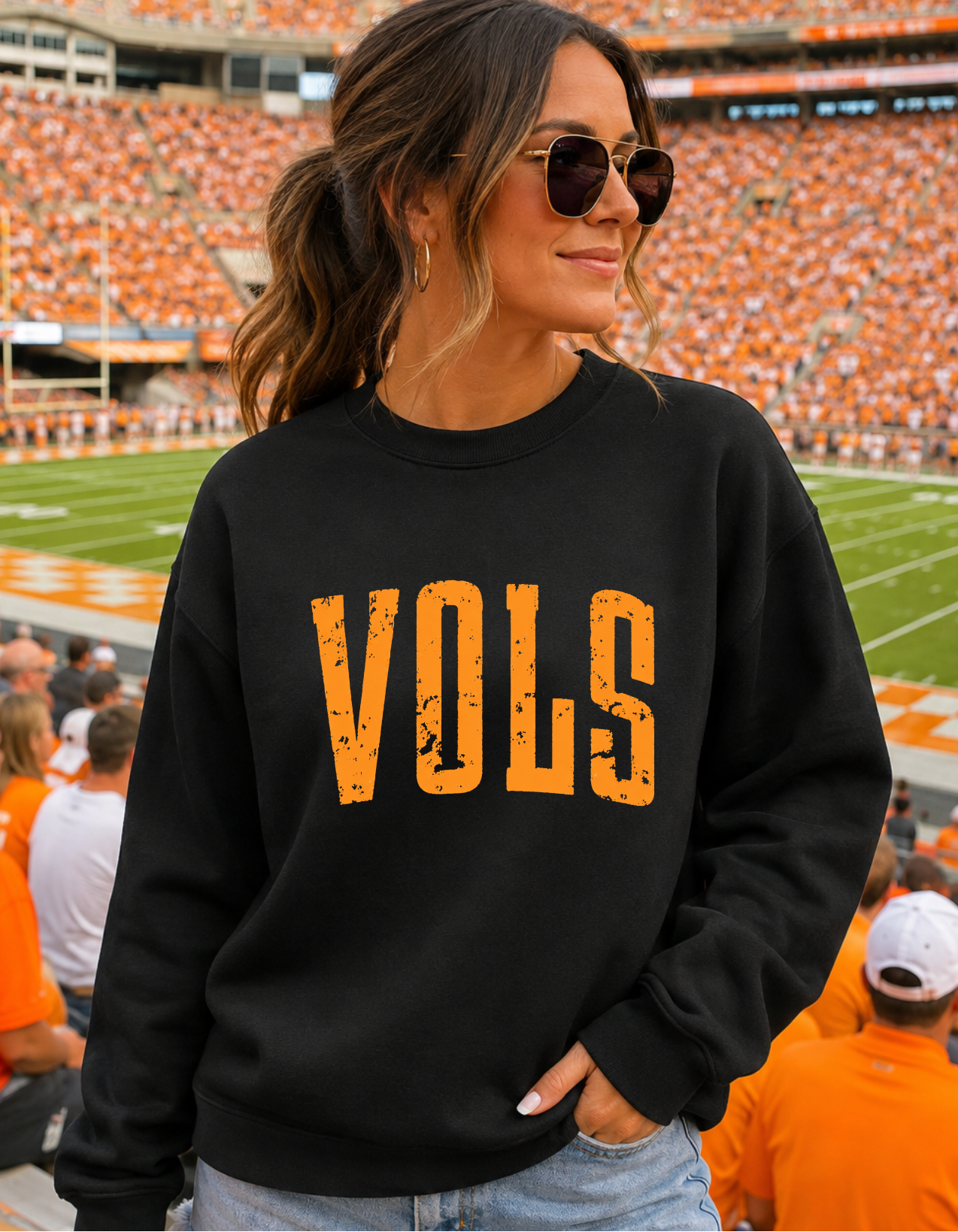 A woman wearing a black crewneck sweatshirt with 'VOLS' printed in distressed orange letters, standing in a stadium with orange seats in the background.