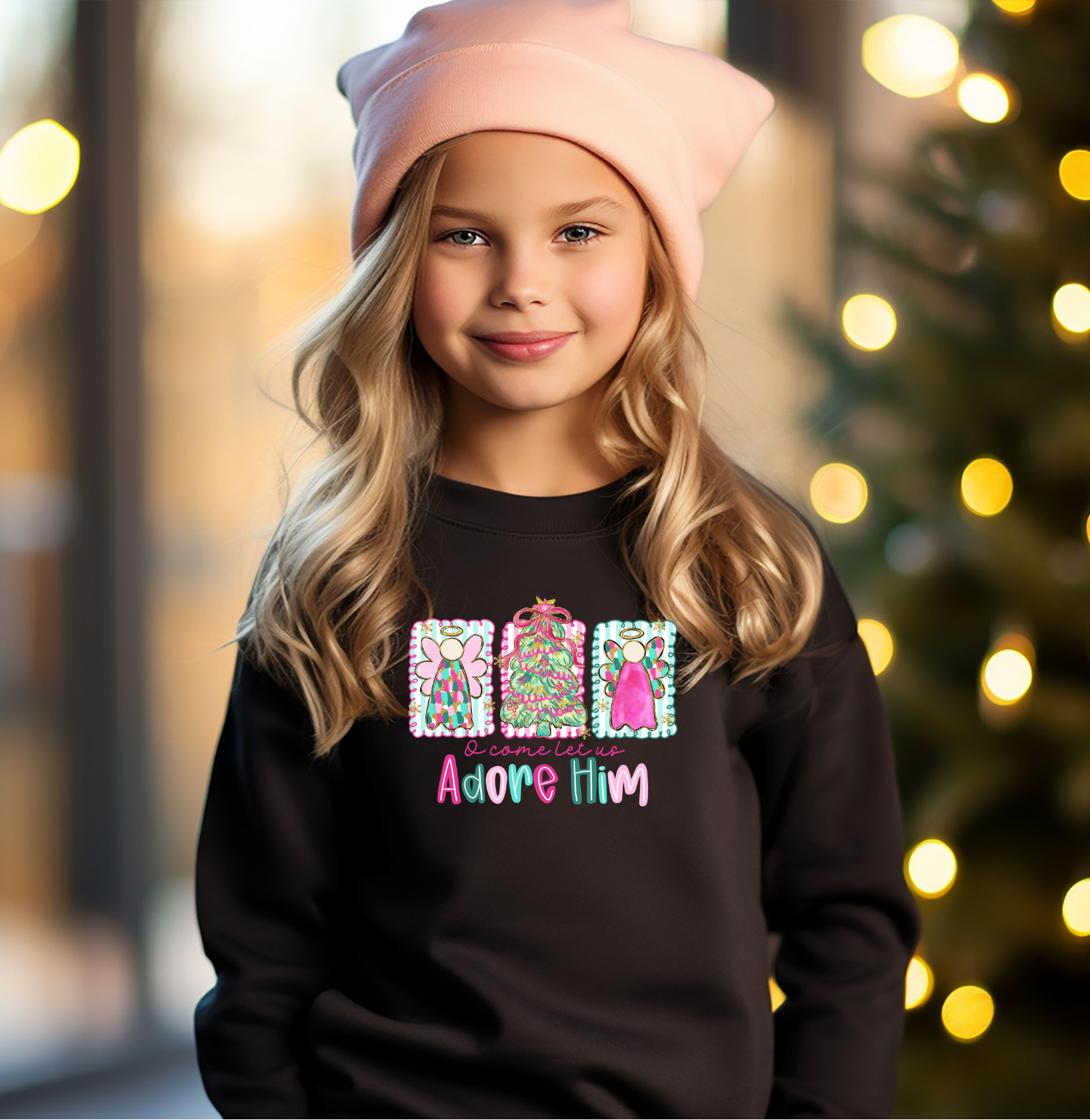 A young girl wearing a black sweatshirt with a bright “O Come Let Us Adore Him” Christmas design, standing near a decorated tree with soft holiday lights in the background.
