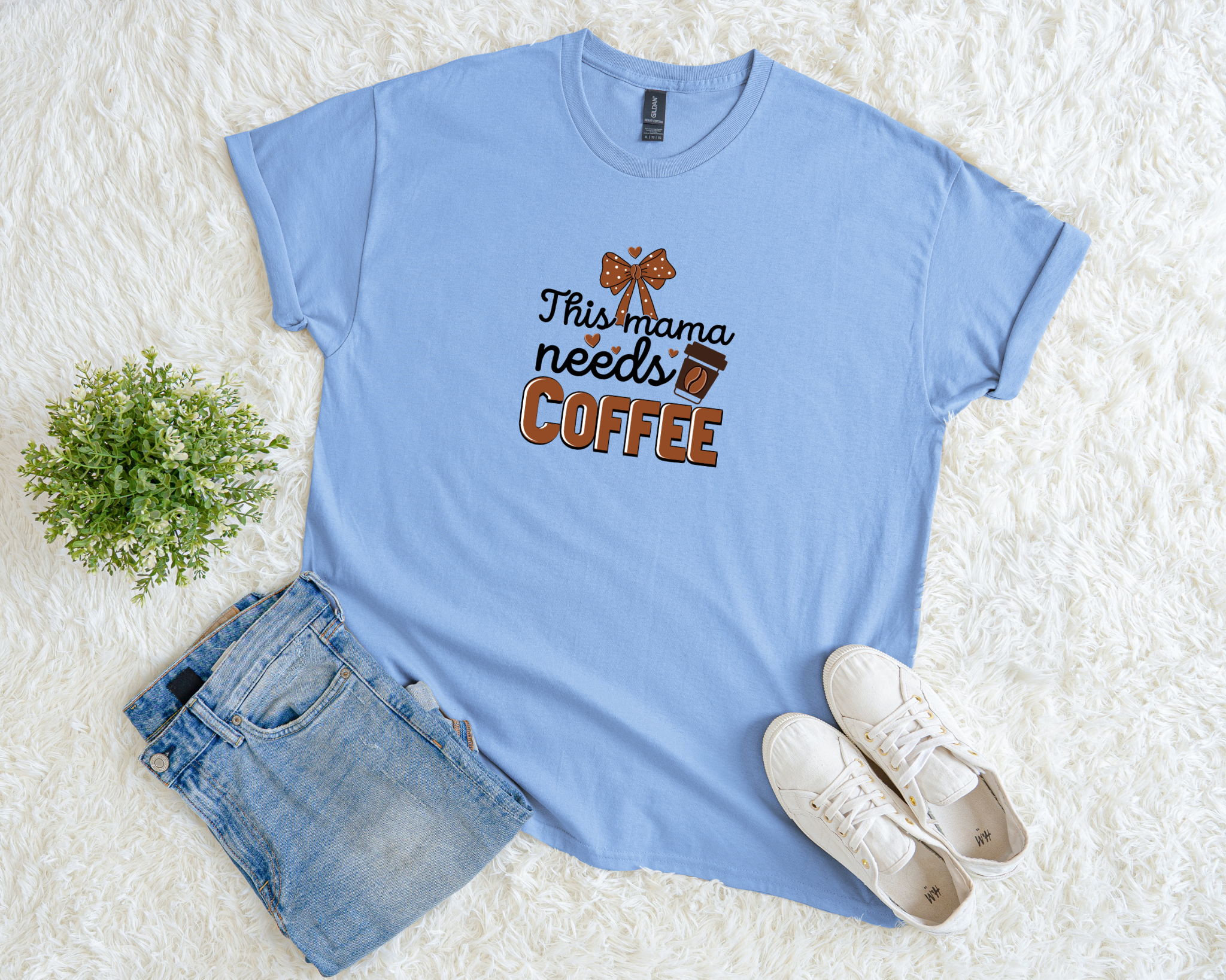 Light blue t-shirt with 'This mama needs coffee' text, paired with denim shorts and white sneakers on a textured surface.