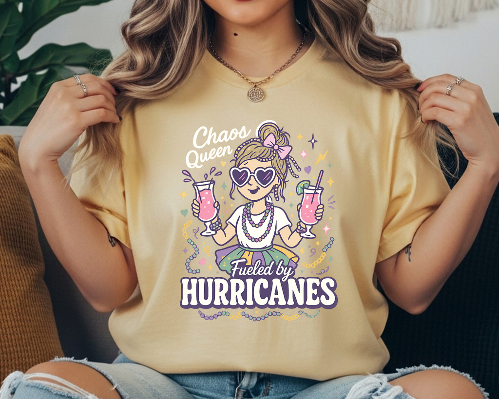 Comfort Colors Mardi Gras t-shirt with a playful girl holding two pink hurricane drinks, surrounded by beads and sparkles, and the text “Chaos Queen Fueled by Hurricanes