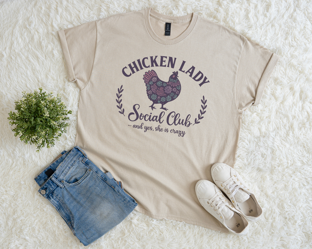 Beige t-shirt with 'Chicken Lady Social Club' graphic on a white surface with denim shorts and sneakers.