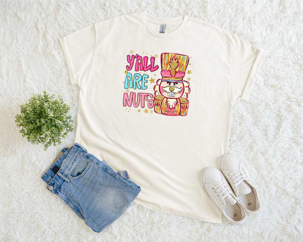 A cream-colored Christmas t-shirt featuring a bright nutcracker design with the phrase “Y’all Are Nuts,” styled flat with jeans and sneakers on a soft white background.