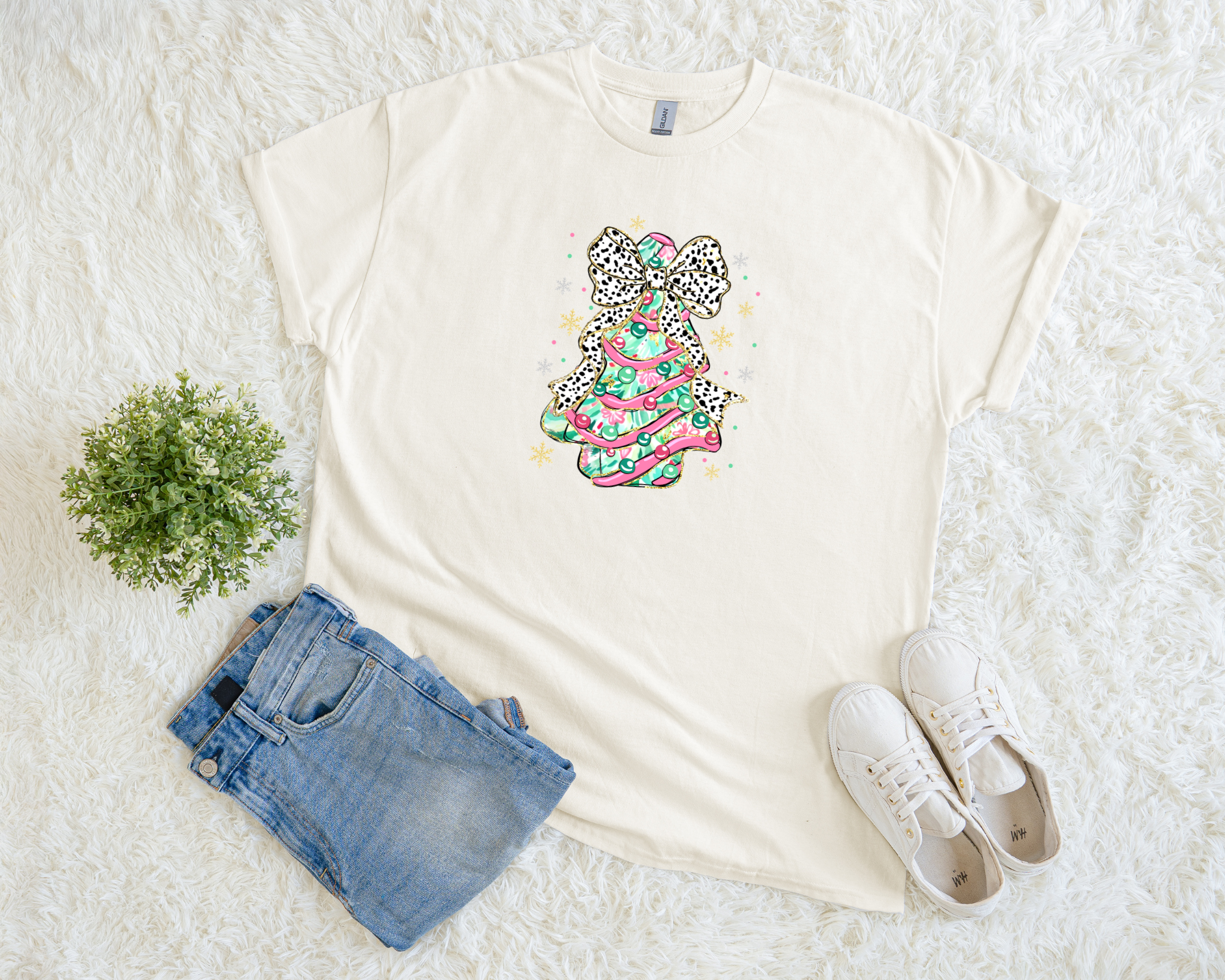 A cream-colored Christmas t-shirt featuring a colorful pink and mint tree topped with a leopard bow, styled flat with jeans and sneakers on a soft white background.
