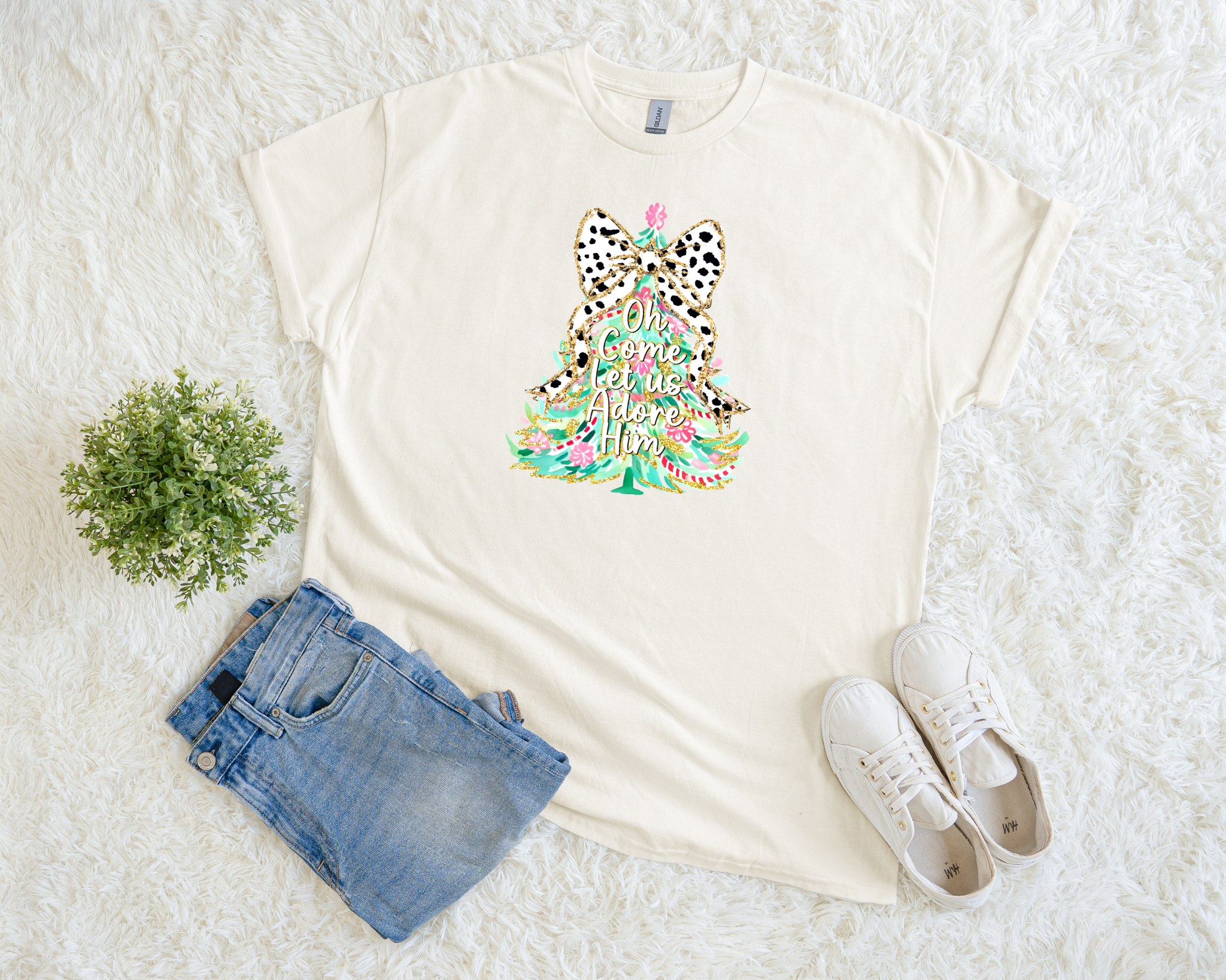 A cream-colored Christmas t-shirt featuring a colorful tree with a leopard bow and the phrase “Oh Come Let Us Adore Him,” styled flat with jeans and sneakers on a soft white background.