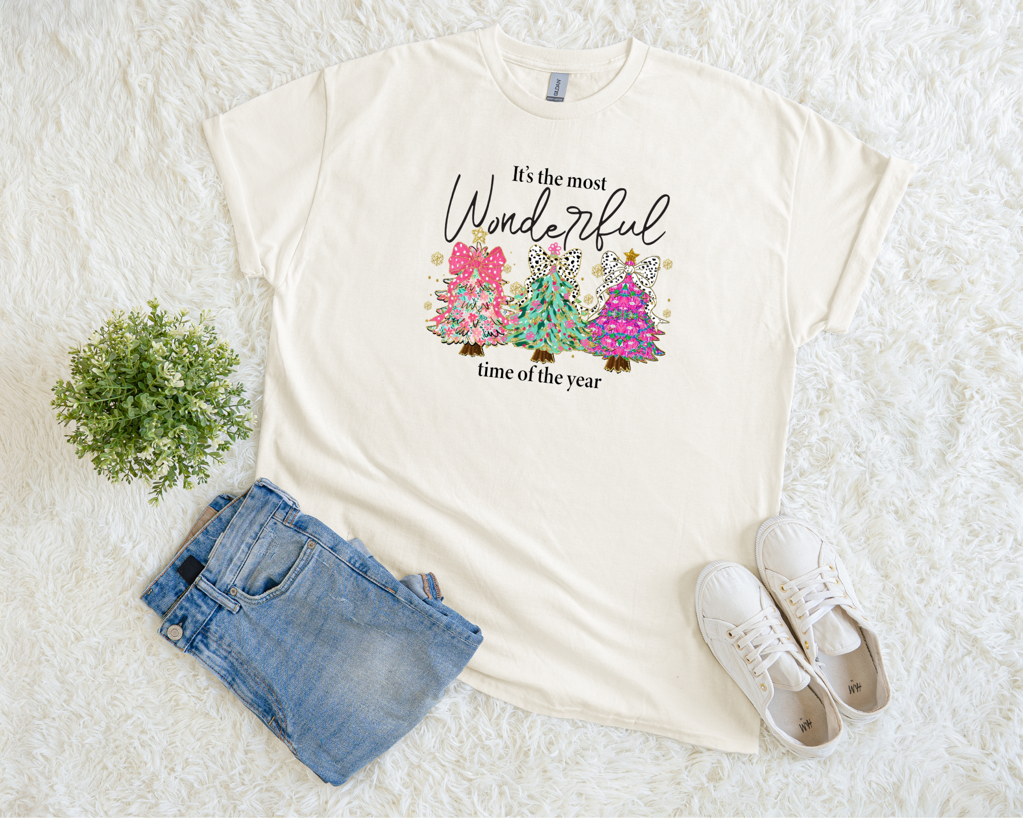 A cream-colored Christmas t-shirt featuring three colorful trees with leopard bows and the phrase “It’s the most wonderful time of the year,” styled flat with jeans and sneakers on a soft white background.