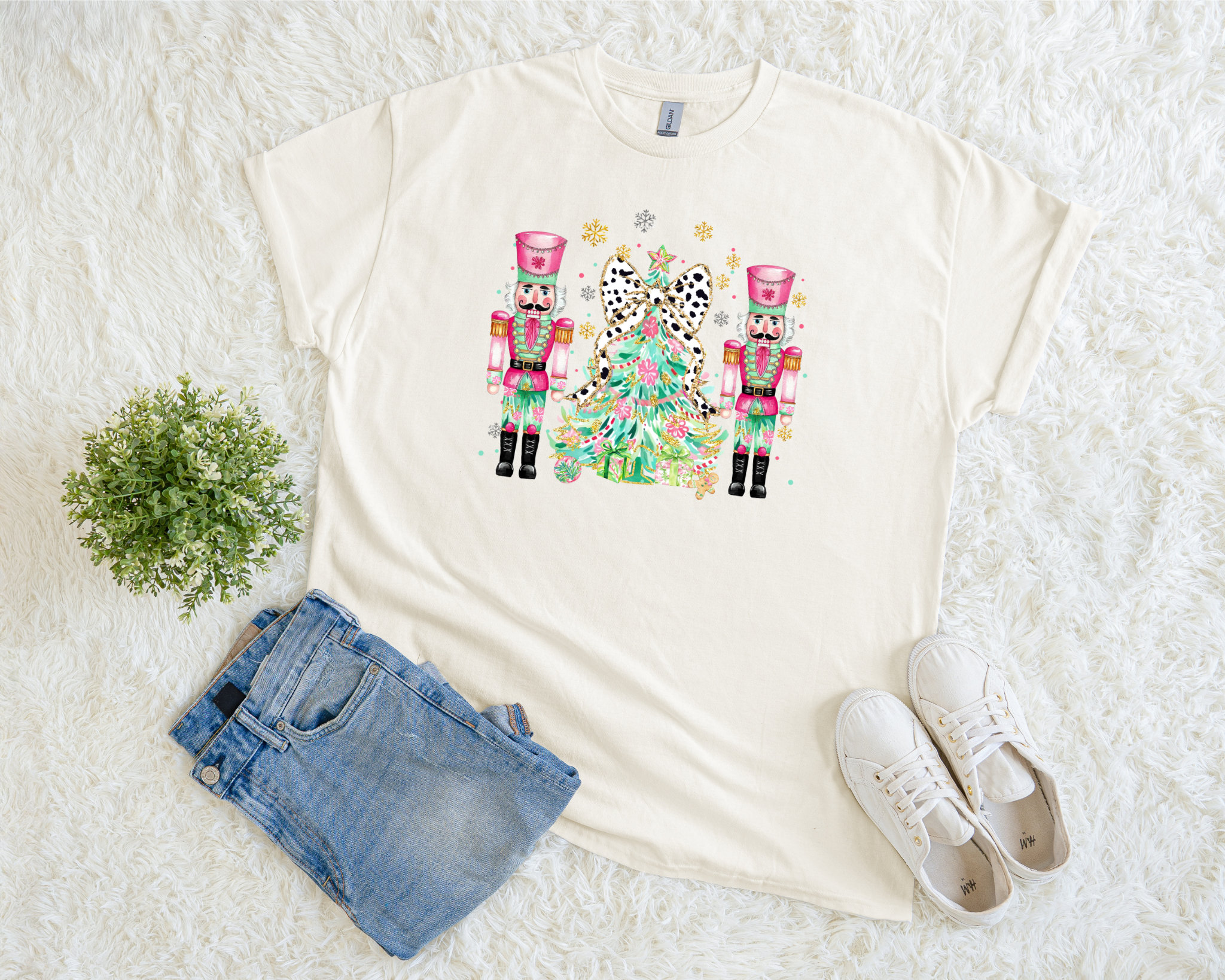 A cream-colored Christmas t-shirt featuring two pink nutcrackers and a leopard bow tree surrounded by snowflakes, styled flat with jeans and sneakers on a white fuzzy background.