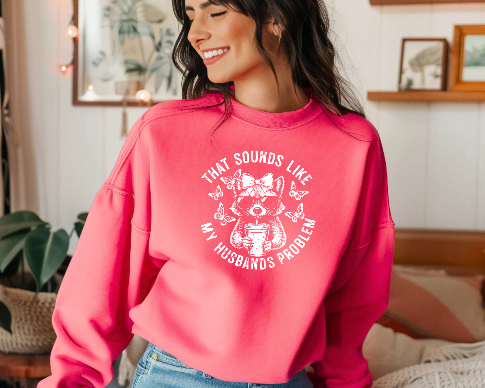 crewneck sweatshirt with white circular graphic of a bow-wearing raccoon in sunglasses holding iced coffee, surrounded by butterflies, reading “That Sounds Like My Husband’s Problem.”