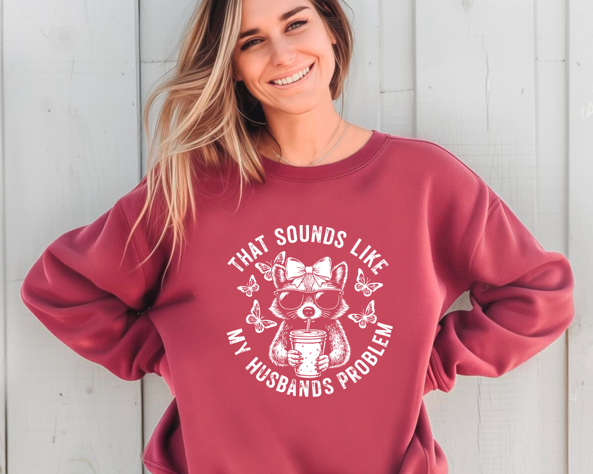 CRIMSON crewneck sweatshirt with white circular graphic of a bow-wearing raccoon in sunglasses holding iced coffee, surrounded by butterflies, reading “That Sounds Like My Husband’s Problem.”