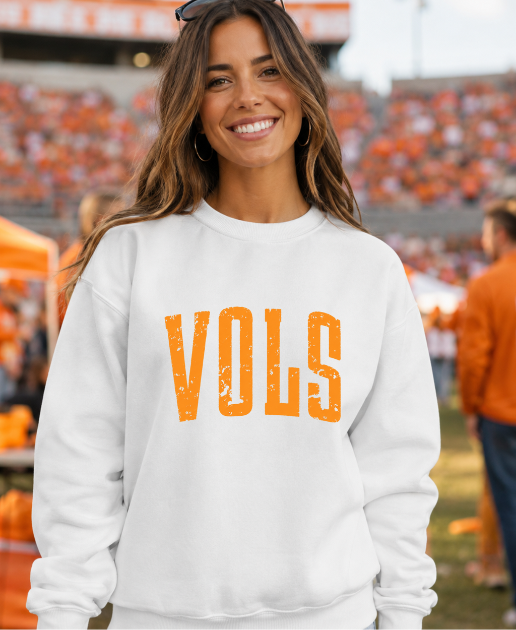 A smiling woman wearing a white crewneck sweatshirt with 'VOLS' printed in distressed orange letters across the chest, standing in a stadium with orange seating in the background.