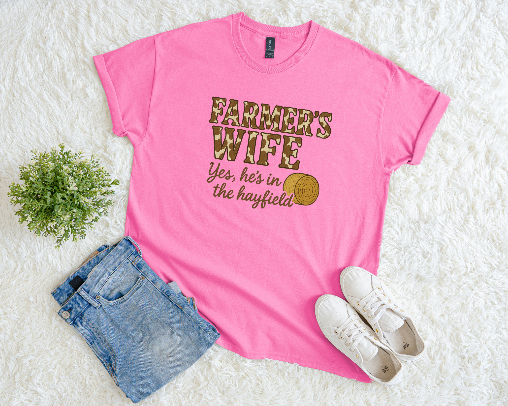 Pink t-shirt with 'Farmer's Wife' text, jeans, and sneakers on a white surface.