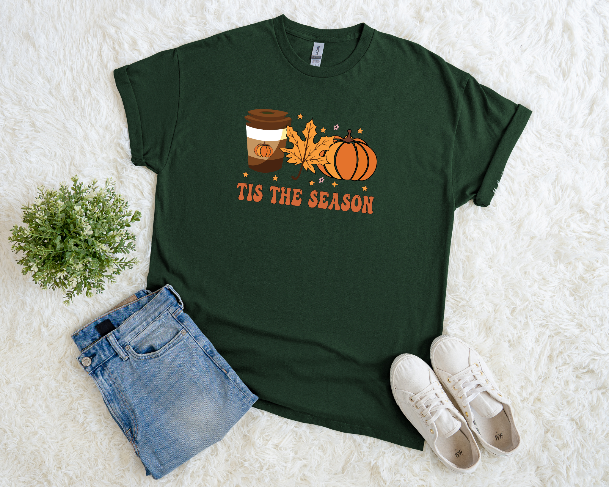 Green t-shirt with 'Tis the Season' design, jeans, and sneakers on a white surface